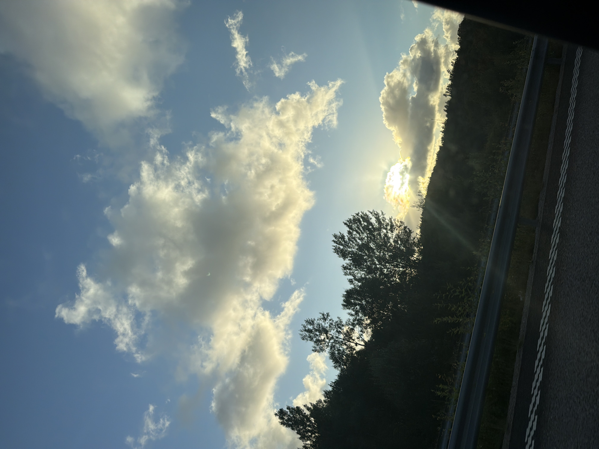 The image shows a view from a car, looking out towards a hillside covered in trees. The sun is low in the sky, partially obscured by clouds, casting a warm glow. There is a guardrail visible in the foreground, separating the road from the hillside. The road itself has double white dashed lines, indicating a lane separation. The sky is blue with scattered, fluffy clouds. The overall scene suggests a scenic drive, possibly during sunset, in or near Ödsmål, Sweden, given the natural landscape and vegetation.