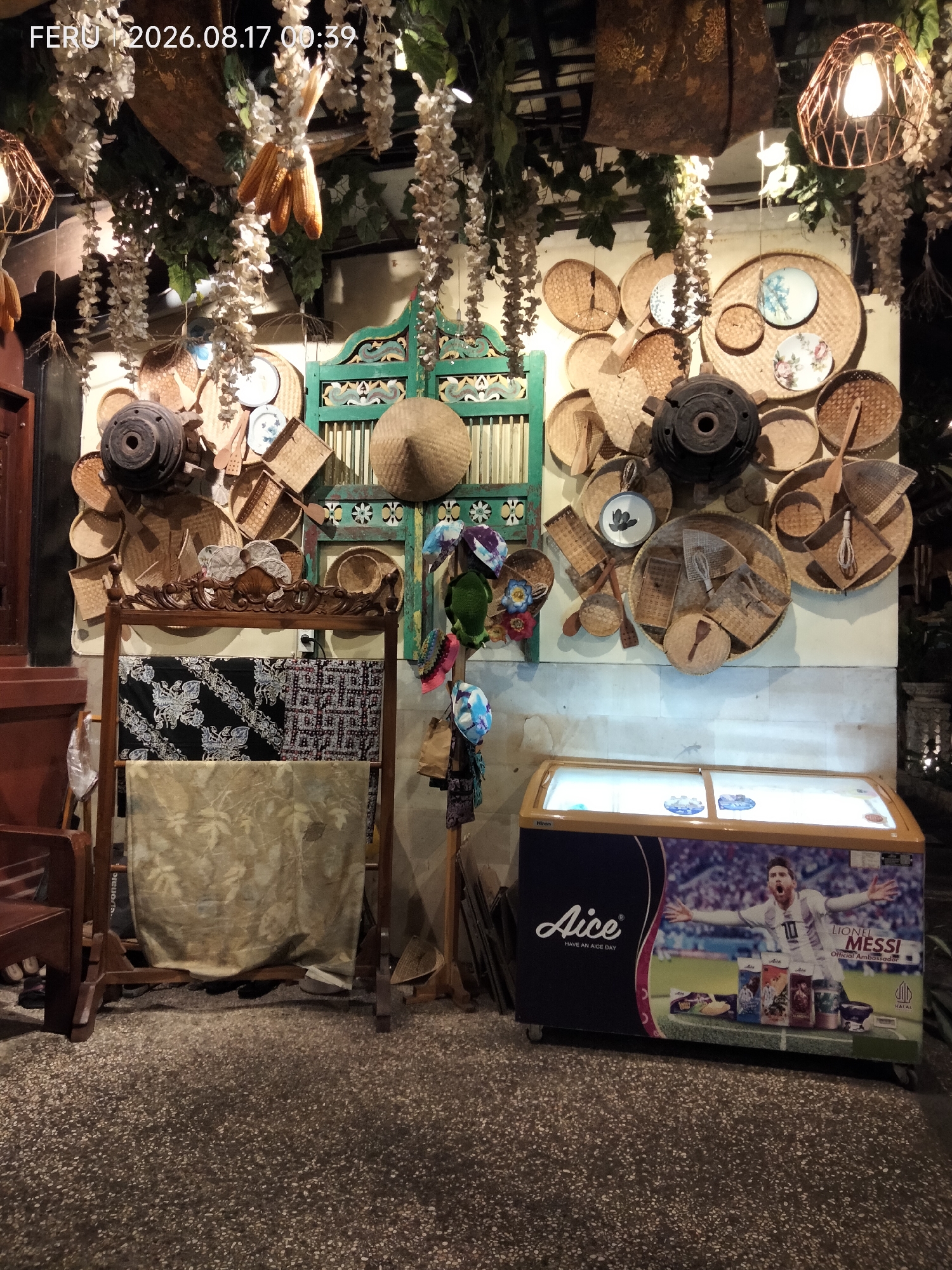 The image depicts a display of traditional Indonesian crafts and products, likely in an outdoor setting or a shop. The central focus is a wall adorned with various woven baskets, plates, and other decorative items made of natural materials. A prominent dark, cylindrical object, possibly a piece of pottery or a decorative element, is centrally placed among these items. To the left of this display, there's a wooden rack holding two pieces of fabric, one black with white intricate patterns and another lighter colored fabric with a leaf motif. Further to the left, a portion of a dark wooden chair is visible. In the foreground, a white and yellow chest freezer is present, featuring an advertisement for "Aice" ice cream with an image of Lionel Messi on its side. The overall atmosphere suggests a cultural or commercial space showcasing local artistry. The timestamp "FERU 2026.08.17 00:39" suggests the image was taken at this date and time. The location is identified as Sukodono, Indonesia.