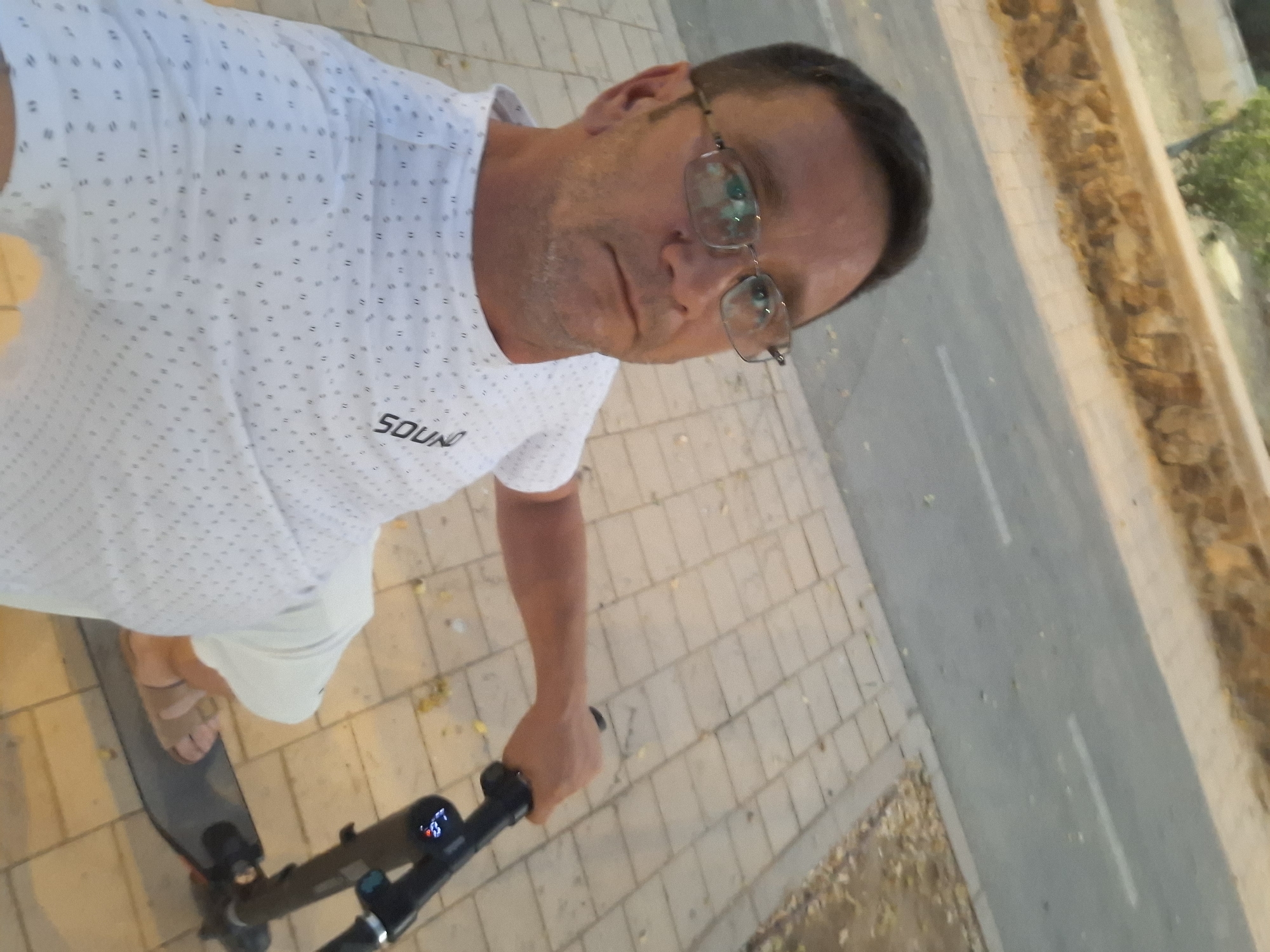 A man with short brown hair and glasses is taking a selfie on an electric scooter on a paved path in Yerocham, Israel. He is wearing a white t-shirt with a repeating pattern of small dark dots and the word "SOUND" in black lettering on the left side. The scooter is black with a digital display showing "1.3". The background shows a road with white markings, a stone wall, and scattered dry leaves on the ground. The lighting suggests it is either early morning or late evening.