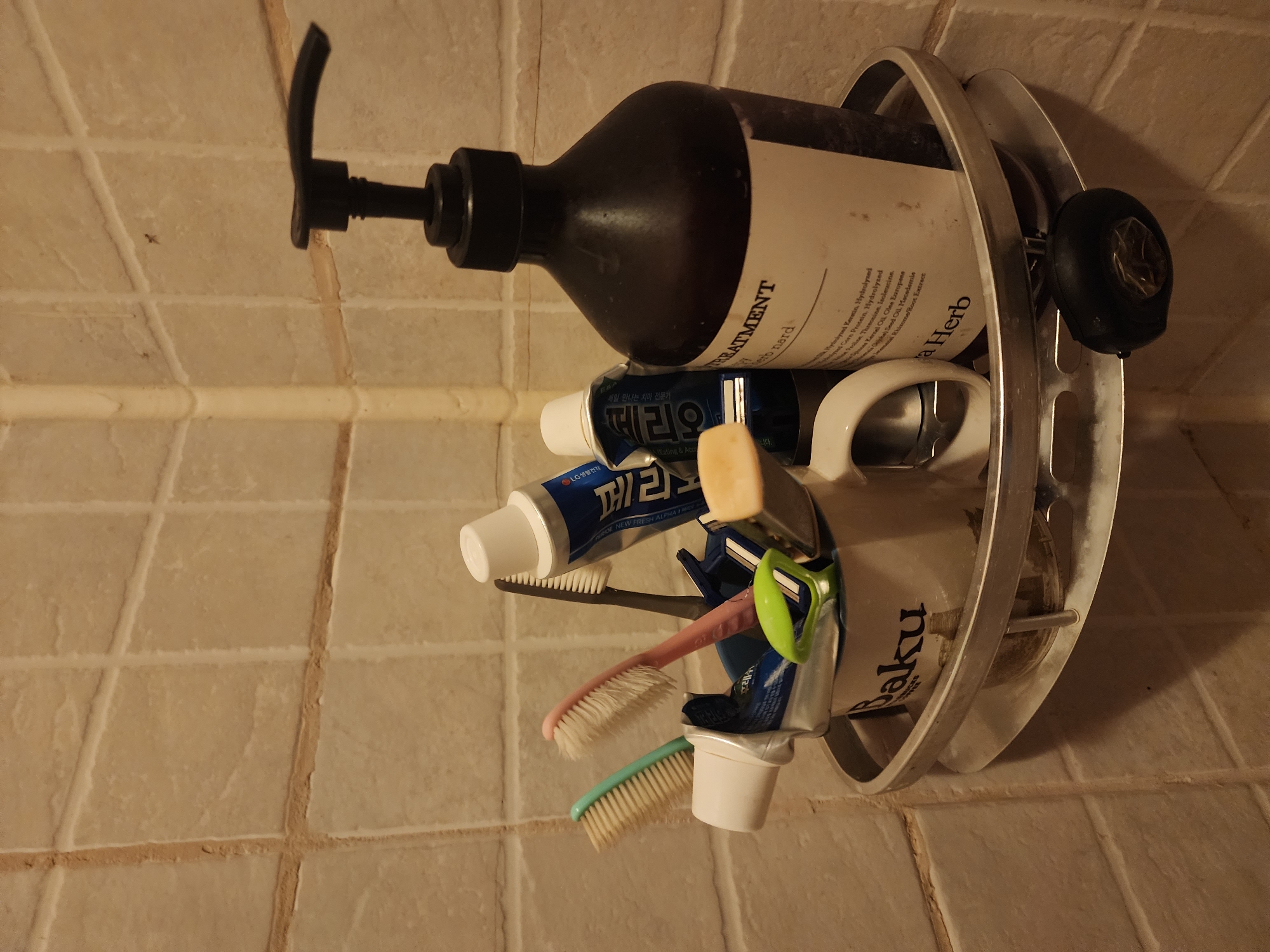 This image depicts a bathroom corner shelf containing various personal hygiene items. The shelf is made of metal with a curved edge and is attached to a beige tiled wall. In the center of the shelf, a white mug with the text "Baku" is visible, holding several toothbrushes and tubes of toothpaste. Among the toothbrushes, there are two with light-colored bristles (one green, one pink), and a dark-colored one. Two tubes of toothpaste are present, one blue and white with Korean text, and another partially visible white tube. A small, light-colored object, possibly a soap bar or a holder, is also on the shelf. To the right of the mug is a large, dark brown pump bottle with a white label. The label has text that appears to include "HERB" and a list of ingredients. The overall environment suggests a typical domestic bathroom setting. The location is identified as Pyeongtaek-si, South Korea, though no specific geographical markers within the image confirm this. There are no people or animals present in the image.
