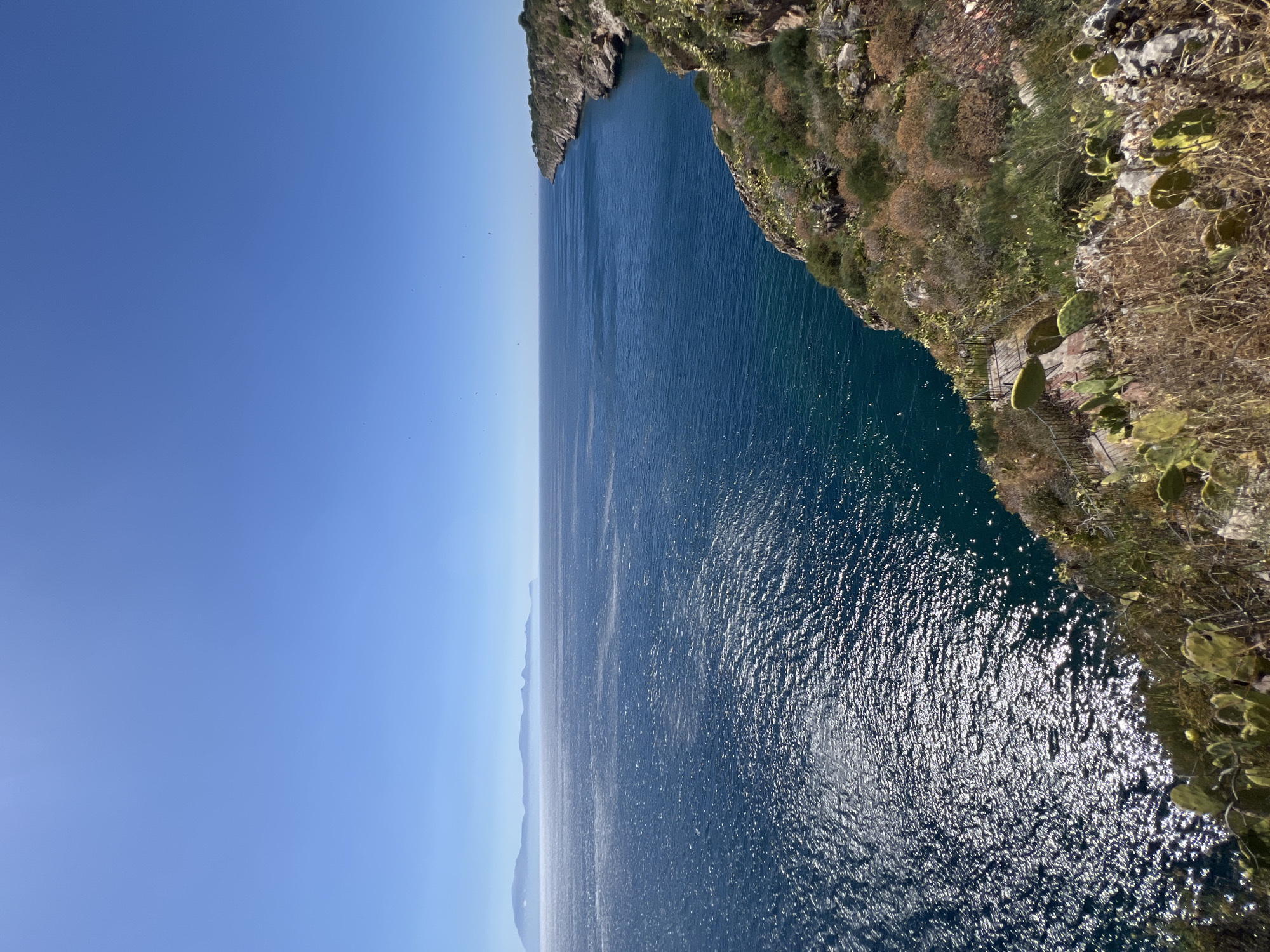 An aerial shot captures the coastline of Terrasini, Italy, with the Mediterranean Sea stretching out to the horizon. The clear blue sky is dotted with a few distant birds. The sea, a deep azure, is illuminated by the sun, creating a shimmering effect on its surface. On the right, a rugged cliff face, covered in dry grass and prickly pear cacti, slopes down towards the water. A winding path with metal railings descends the cliff, suggesting a scenic overlook or trail. In the distance, hazy mountains form a silhouette against the horizon. The scene is peaceful and serene, evoking a sense of natural beauty and tranquility.