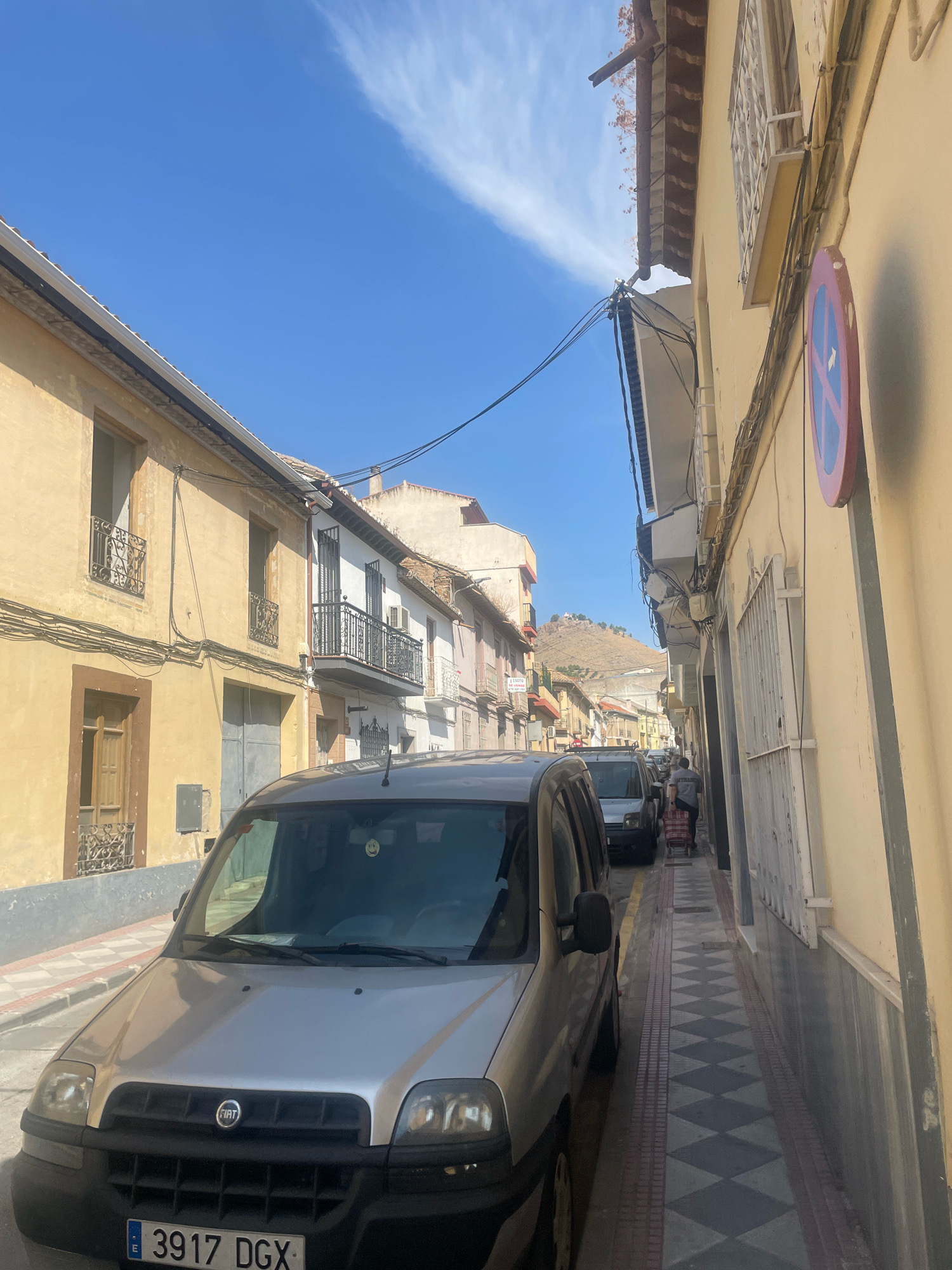 A street scene in Atarfe, Spain, depicts a narrow, sunlit alleyway flanked by buildings. In the foreground, the front of a silver Fiat van with license plate "3917 DGX" is visible. The van is parked on the left side of the street, which has a sidewalk with a diamond-patterned tile design in grey and red.
The buildings lining the street are constructed in a traditional style, with yellow and white facades, balconies with ornate metal railings, and visible windows. Overhead, power lines crisscross the sky. Further down the street, more buildings are visible, some appearing older and less maintained. In the distance, a hillside with some structures is present.
A person is seen walking away from the camera down the sidewalk, pulling a red and white striped shopping cart. Another vehicle, possibly a van, is parked in front of them. A circular road sign with a red border and a blue background with a white arrow pointing to the right is mounted on the wall of a building on the right side of the street. The sky is bright blue with some wispy white clouds. The overall impression is a typical urban street scene on a clear day.