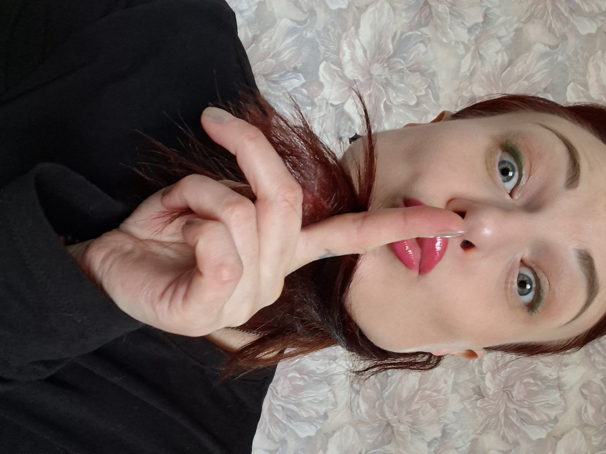 A woman with red hair is lying on a white floral patterned surface. She is wearing a black top and has her index finger to her lips in a "shhh" gesture. Her lips are painted with a glossy pink lipstick. She has green eyeshadow on her eyelids. A chain is visible near her neck. The location is Забалоцкі сельскі Савет, Belarus.