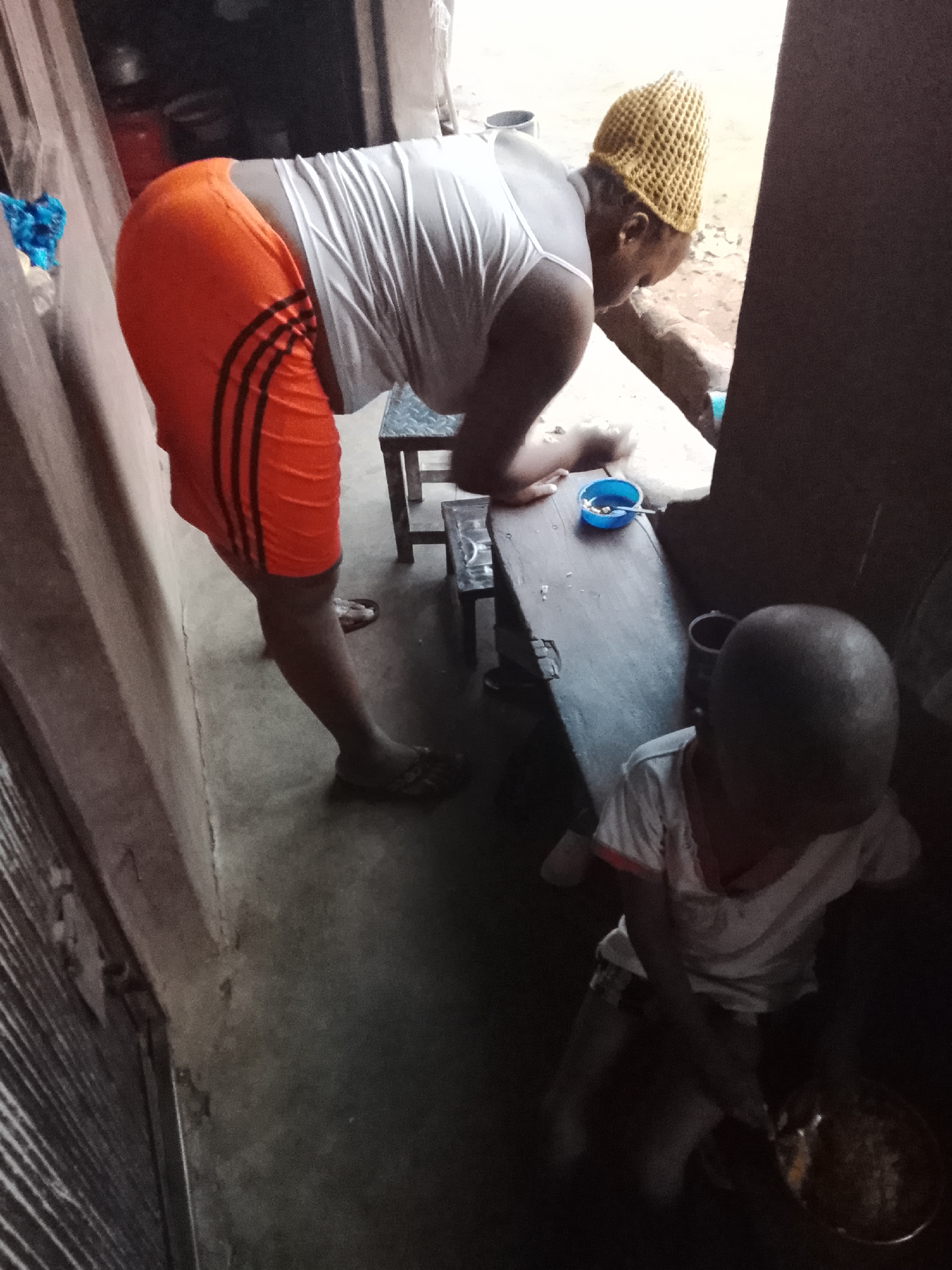 This image captures a domestic scene, likely during the daytime in Nkpor, Nigeria, given the natural light source from an opening behind the main subject. In the foreground-midground, a dark-skinned woman is bent over, engaged in a task. She is wearing bright orange athletic shorts with three black stripes down the side, a white sleeveless top, and a distinctive yellow knitted or crocheted hat. Her feet, clad in flip-flops, rest on a concrete floor. She appears to be leaning over a low wooden surface where a small blue bowl, possibly containing food, is placed.
Further into the background and to the right, a young child with short, dark hair is seated on the floor or a very low stool. The child is wearing a white t-shirt and appears to be occupied with a small pot or pan. The setting is simple, with concrete flooring and rustic or unadorned walls. Various household items and containers are faintly visible in the somewhat dim background. The overall activity suggests daily life, possibly involving food preparation or a shared meal within a home environment. There is no visible text in the image.