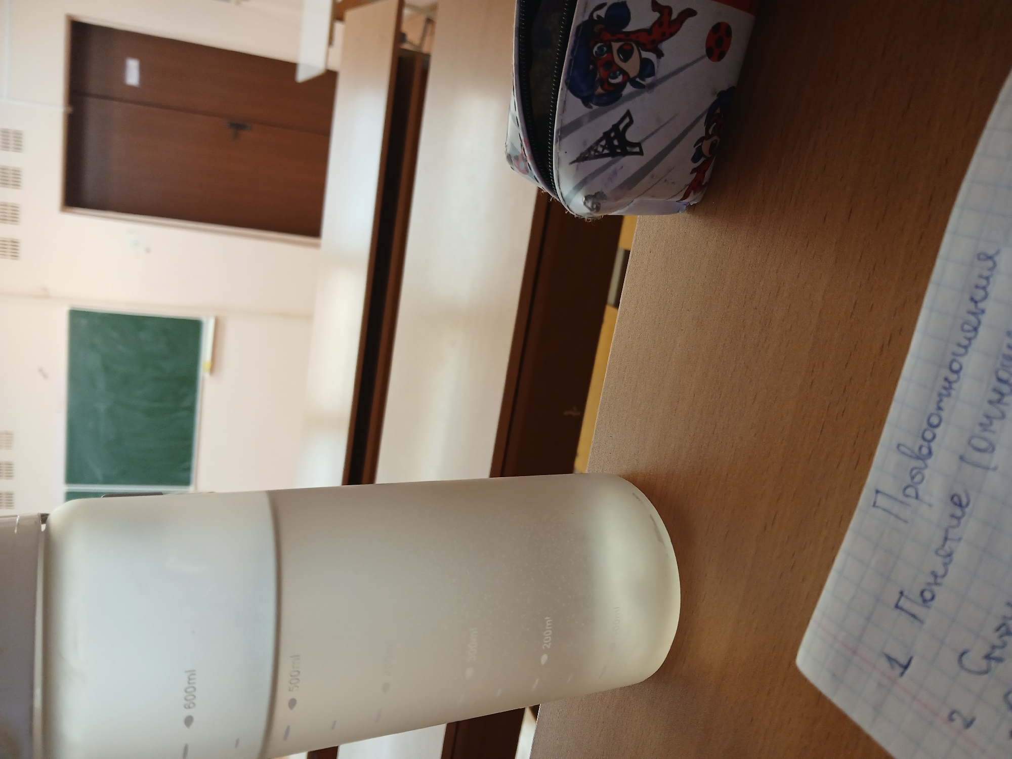 The image shows a desk in a classroom setting, likely in Chaikovsky, Russia based on the Cyrillic handwriting on a notebook. A water bottle with volume markings (600ml, 500ml, 300ml, 200ml) is positioned horizontally. Next to it is a notebook with handwritten notes in Russian. Adjacent to the notebook is a pencil case with a Ladybug design. Background elements include a brown door, a green chalkboard, and empty desks/seats.