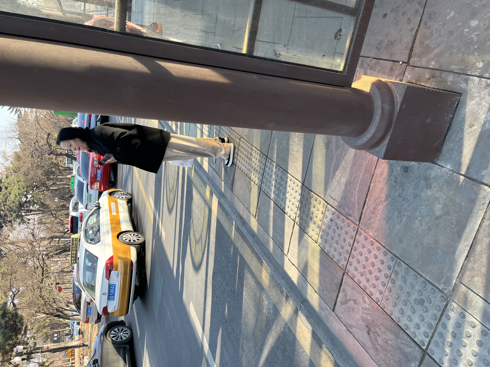 The image captures a street scene in Changchun City, China. An Asian woman with black hair, wearing a black coat and light-colored pants, stands on the sidewalk near a bus shelter or building with a large column. She is holding a cell phone. A taxi with a white and gold color scheme and license plate "吉Y4842" is parked parallel to the sidewalk. There are several other cars parked behind the taxi. The background shows trees without leaves. The sidewalk is paved with gray stone tiles and tactile paving for the visually impaired. Shadows from the bus shelter and trees are cast onto the pavement.
