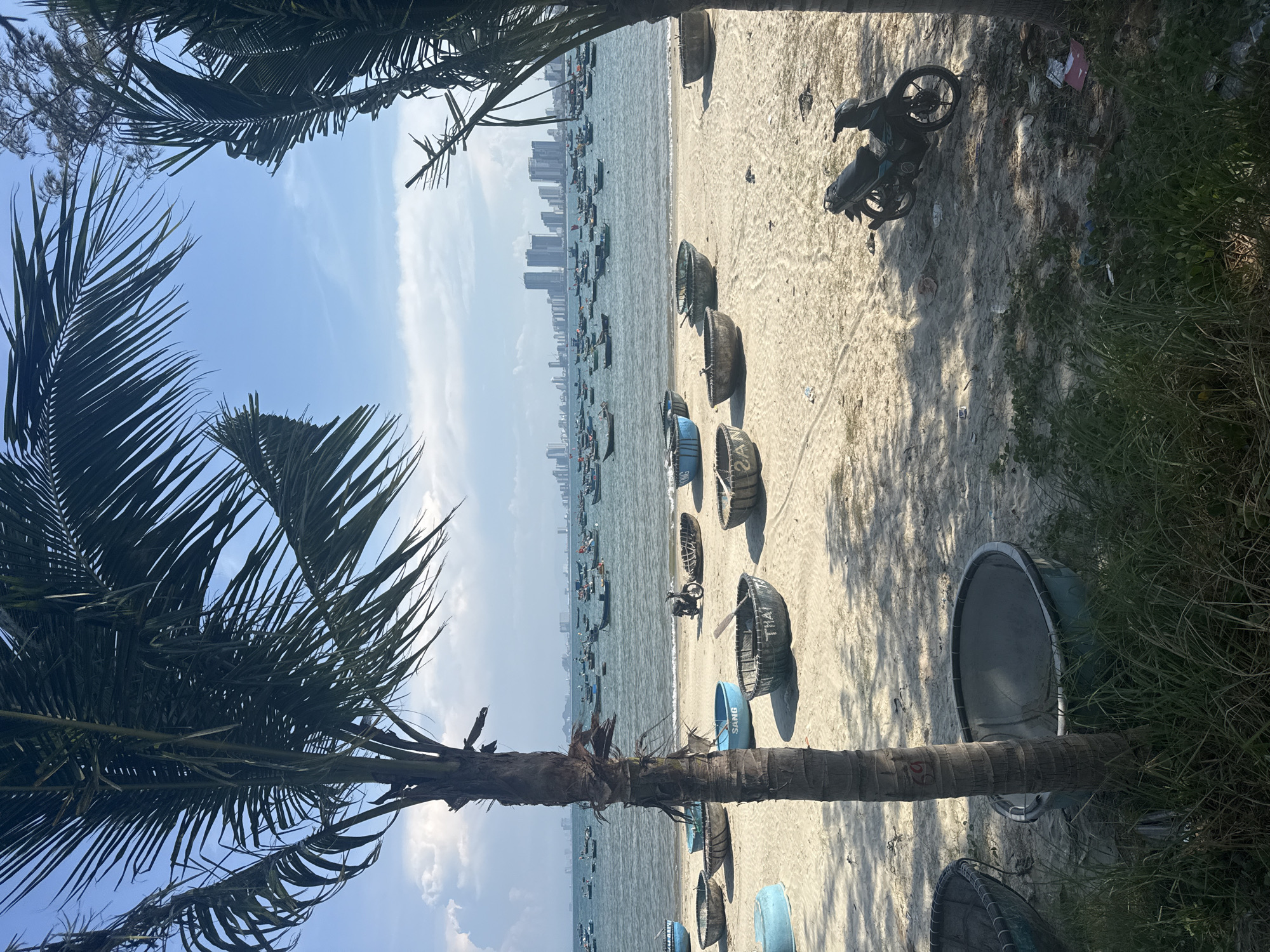 The image depicts a beach scene in an unidentified city. Palm trees frame the left side of the image, obscuring the sky with their fronds. In the distance, a city skyline is visible. The beach is lined with numerous traditional fishing boats, some of which are blue and others a dark color. A motorcycle is parked on the sand near the upper right corner. Grass and other vegetation grow along the edge of the beach on the right side of the image.