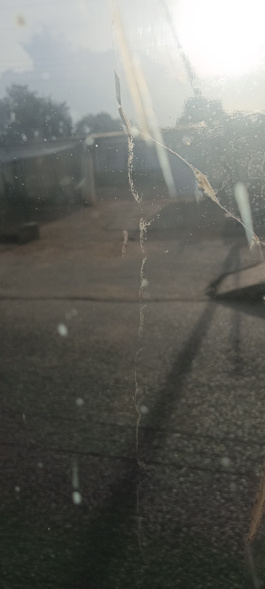 The image captures an outdoor scene viewed through a dirty and potentially scratched or cracked glass panel. The foreground is dominated by the semi-transparent surface, which is heavily obscured by smudges, dust particles, and various marks. A prominent vertical line of grime or a thin crack runs down the right-center of the glass, while other finer scratches and water spots are scattered across its surface.
Through the textured glass, a blurry and hazy background is visible. Shapes indicative of trees or foliage can be discerned on the left and center, suggesting a natural or vegetated outdoor environment. In the upper right, a bright, diffused light source, likely the sun, creates significant glare and reflections on the glass, further obscuring the distant view. This strong light suggests either morning or late afternoon. No specific activities or individuals are visible. The overall impression is of an observer looking out from an enclosed space, with the immediate barrier severely limiting the clarity of the external world. There is no visible text in the image. While the location context is Jos, Nigeria, the image itself does not contain specific visual cues to confirm this.