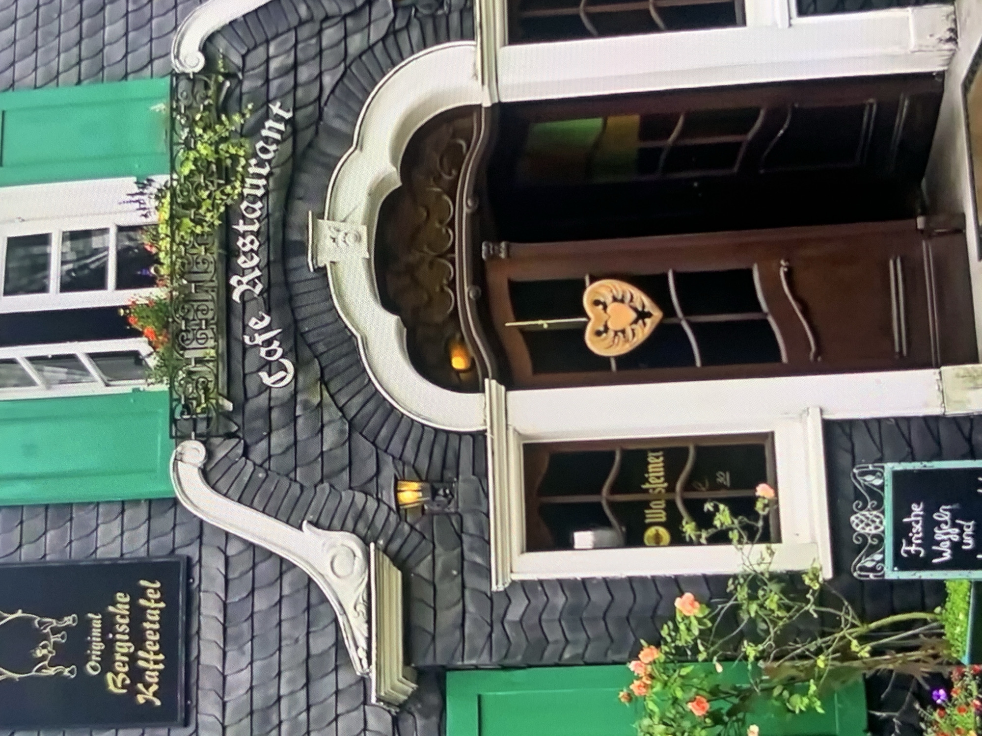 The image shows the exterior of a cafe and restaurant with a distinctive architectural style. The building features a dark, slate-tiled roof with ornate white trim. There are green shutters on the upper windows, and a window box filled with flowers and ivy adorns the facade. A sign above the entrance reads "Cafe Restaurant" in white, Gothic-style lettering. The entrance itself consists of dark wooden double doors, one of which is slightly ajar, revealing a dimly lit interior. A heart-shaped wooden decoration hangs from the door. To the left of the entrance, there is a window with a sign that appears to say "Wa.steiner." Below this window, a small blackboard displays text that includes "Frische Waffeln" (Fresh Waffles). A small tree with pinkish-orange roses is planted to the left of the entrance. The overall impression is that of a charming, traditional establishment. The image does not provide any information about the city of Stavropol, Russia.