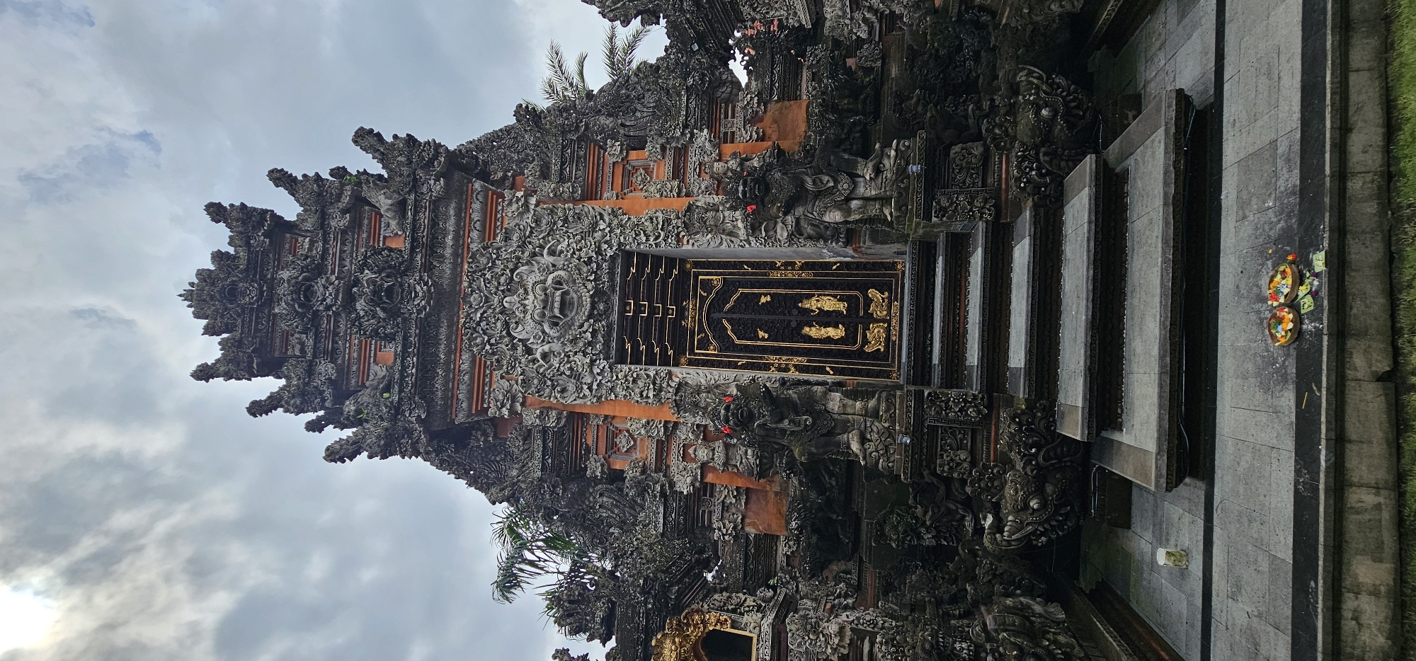 The image captures a temple gate in the Ubud District, Indonesia, featuring intricate stone carvings and decorations. The gate has a black door with gold trim and depictions of figures. Steps lead up to the gate, with offerings of flowers and other items placed at the base of the steps. The sky is overcast, and the surrounding area appears to be a mixture of stone and greenery.