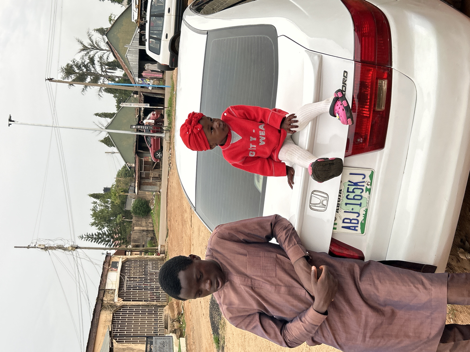 A medium, rotated shot presents a white Honda Accord in an outdoor, urban environment, likely in Jos, Nigeria. A young child in a red sweater and headscarf sits on the car's trunk, legs outstretched. A man stands next to the car, leaning slightly on the trunk near the license plate, which reads "ABJ-165KJ, ABUJA." The background features buildings, utility poles, and a dirt ground.
