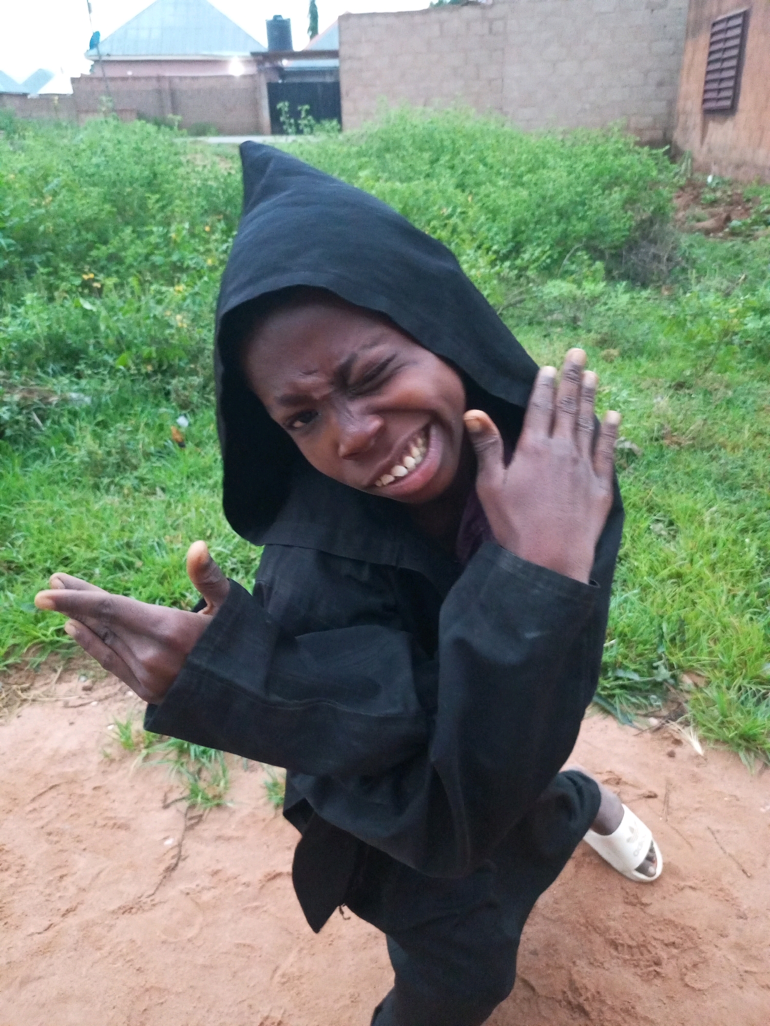 In Tempure, Nigeria, a young Black individual, possibly a child or adolescent, stands outdoors on a dirt path, their face turned towards the camera with a playful expression and a wink. They are wearing a black hooded robe, which obscures most of their body. Their left hand is held out, palm facing forward, while their right hand is raised in a gesture that could be interpreted as a wave or a playful warding off. Behind them, a patch of green grass and shrubs borders a reddish-brown dirt ground. In the background, a low brick wall and part of a building with a metallic roof are visible, suggesting an urban or semi-urban environment. The lighting indicates it is daytime.