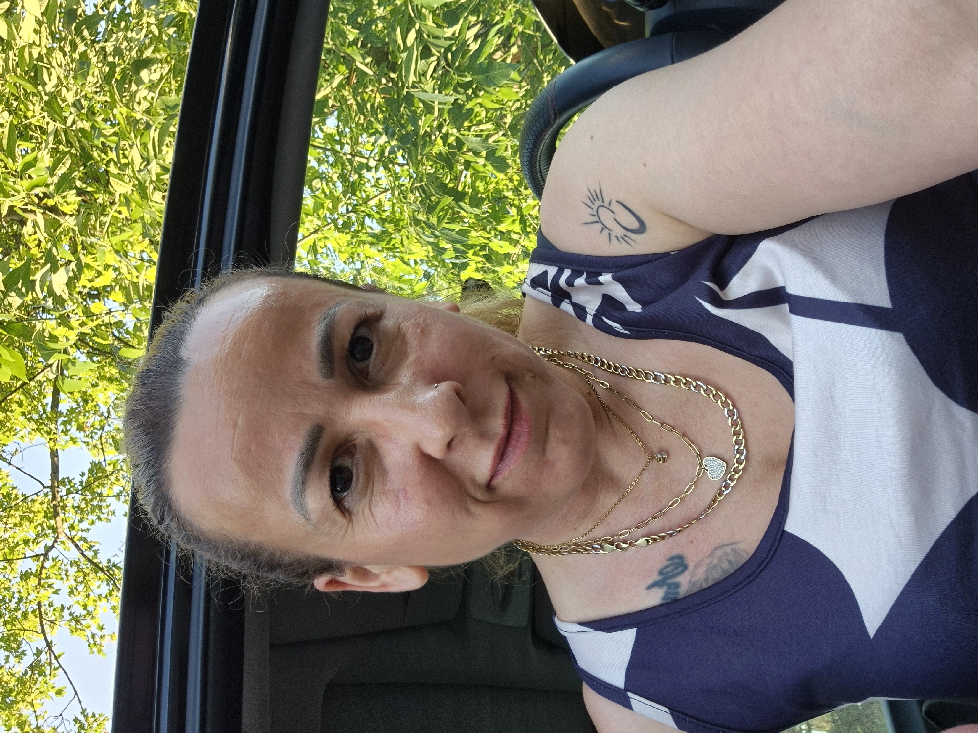 A woman is captured in a close-up, slightly tilted photograph, possibly a selfie. She looks directly at the camera with a subtle smile. Her eyes are dark, and her hair, pulled back, shows a mix of light brown/blonde and grey. A small silver stud pierces her right nostril.
She is wearing a blue and white patterned tank top or dress. Several gold chains adorn her neck, one featuring a small, glittering heart-shaped pendant, and another with a tiny clear stone. On her right shoulder/upper arm, a prominent black tattoo depicts a sun symbol connected to a crescent or 'C' shape. Part of another tattoo is visible on her upper chest, beneath her necklace.
The background suggests she is inside a vehicle, with a dark window frame visible on the left side of the image. Through the window, vibrant green foliage of trees and a bright blue sky are visible, indicating an outdoor, sunny setting during the daytime. The overall impression is casual and relaxed. There is no visible text in the image apart from symbolic tattoos.
