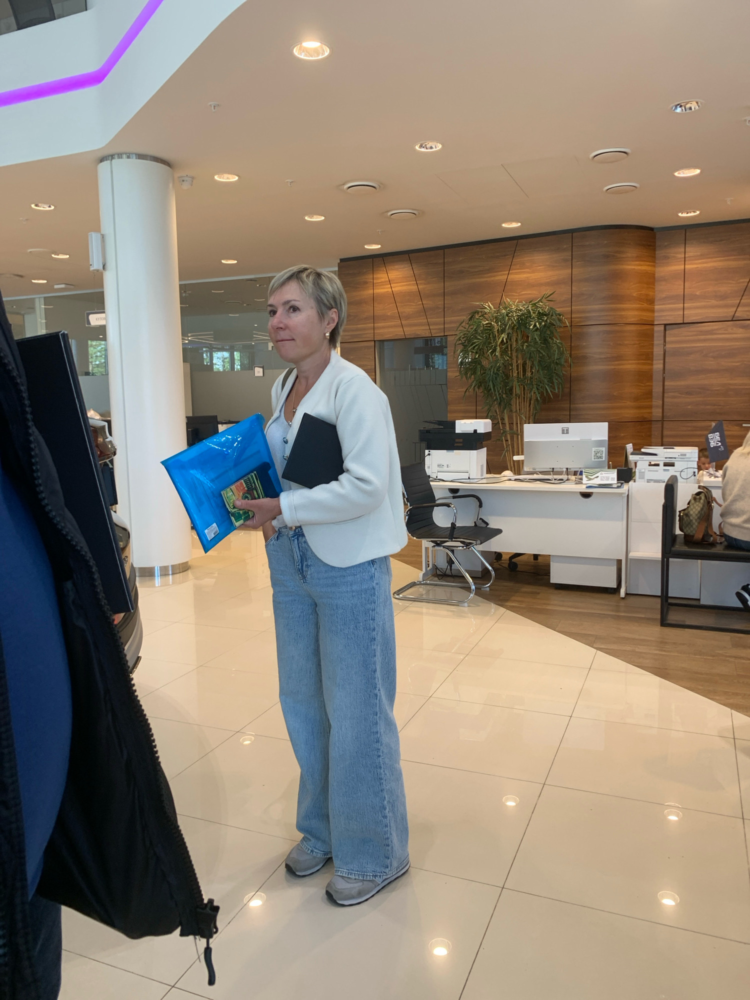 A woman with short blonde hair stands in a brightly lit, modern interior that appears to be a car dealership. She is wearing a white cardigan, a light-colored V-neck top, and loose-fitting light blue jeans. The woman is holding a bright blue plastic folder and a black folder. Her left hand is in the blue folder, and her right hand is holding the black folder. She is looking to her right.
The environment features a tiled floor with embedded lighting, white cylindrical pillars, and a curved wall with wood paneling. In the background, there is a reception desk with computers, printers, and office supplies. A tall potted plant is situated behind the desk. The lighting is a mix of recessed ceiling lights and ambient light reflecting off the polished surfaces. The overall impression is one of a clean, contemporary commercial space. There is no specific indication that this location is in Minsk, Belarus.