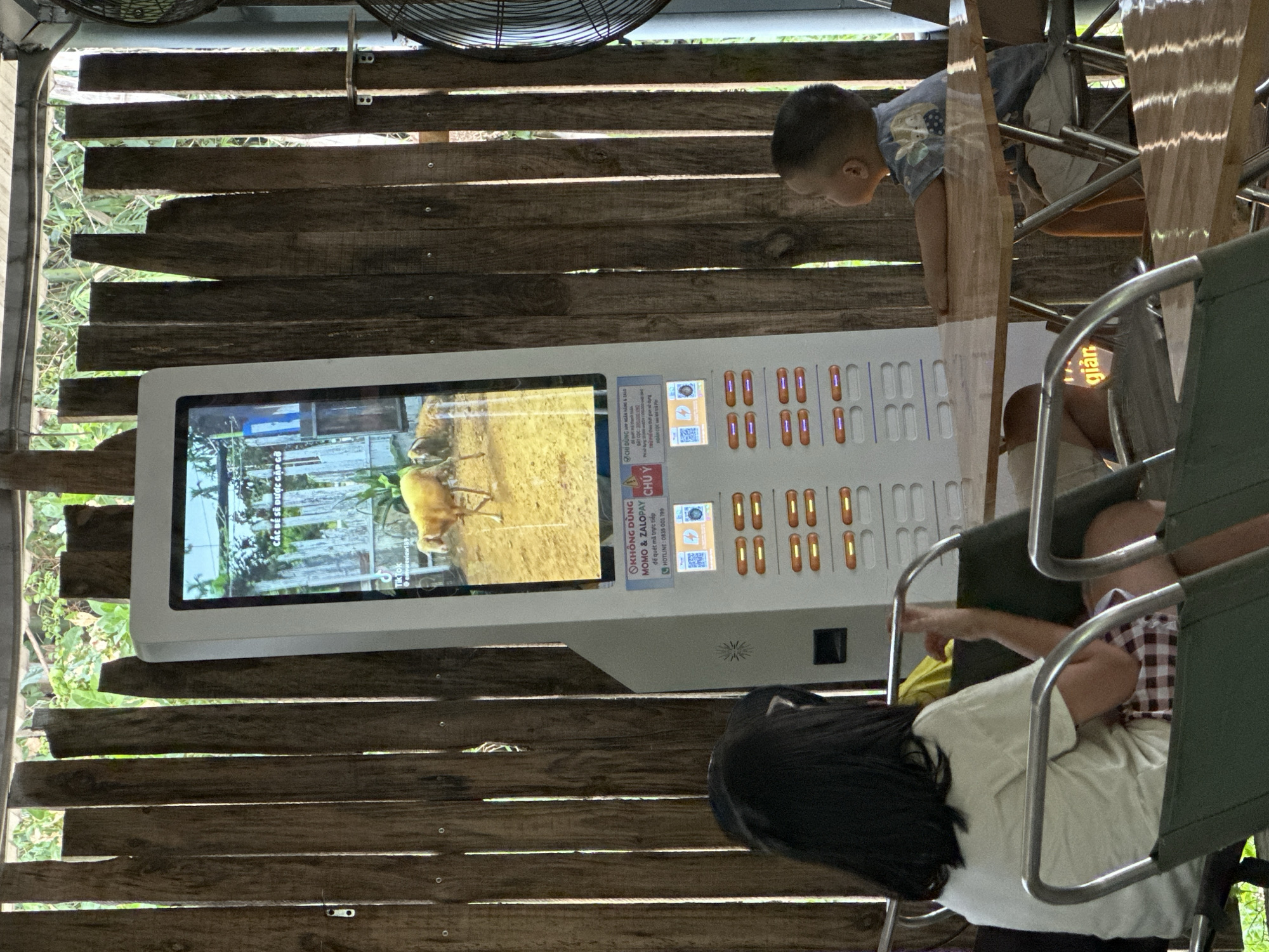 This image captures a scene at what appears to be an outdoor attraction, possibly a zoo or animal park, in Đà Nẵng, Vietnam. The setting is rustic, with weathered wooden planks forming a backdrop and partitions. In the foreground, a modern touch kiosk with a large screen is set up. The screen displays an image of an animal, possibly a deer or antelope, in a naturalistic enclosure. The kiosk also features a series of buttons or selection options, indicated by rows of illuminated rectangular shapes and accompanying text in Vietnamese. Some text on the kiosk provides instructions and contact information, including a hotline number and mentions of payment methods like "MOMO & ZALOPAY" and "APP NGÂN HÀNG & TALO."
Two young children are present in the image. One child, a boy wearing a t-shirt with a cartoonish print, is looking intently at the screen of the kiosk, his head and shoulders visible. He appears engaged with the display. Another child, a girl with dark hair, is partially visible in the foreground, seated or leaning, also seemingly interacting with the kiosk or the environment. The presence of seating with metal frames suggests a waiting or viewing area.
The overall impression is a family-friendly environment, likely during the daytime, given the natural light filtering through the wooden structures. The atmosphere seems relaxed and observational, with the children engrossed in the interactive display. The juxtaposition of rustic natural materials with modern technology is a notable detail of the setting.