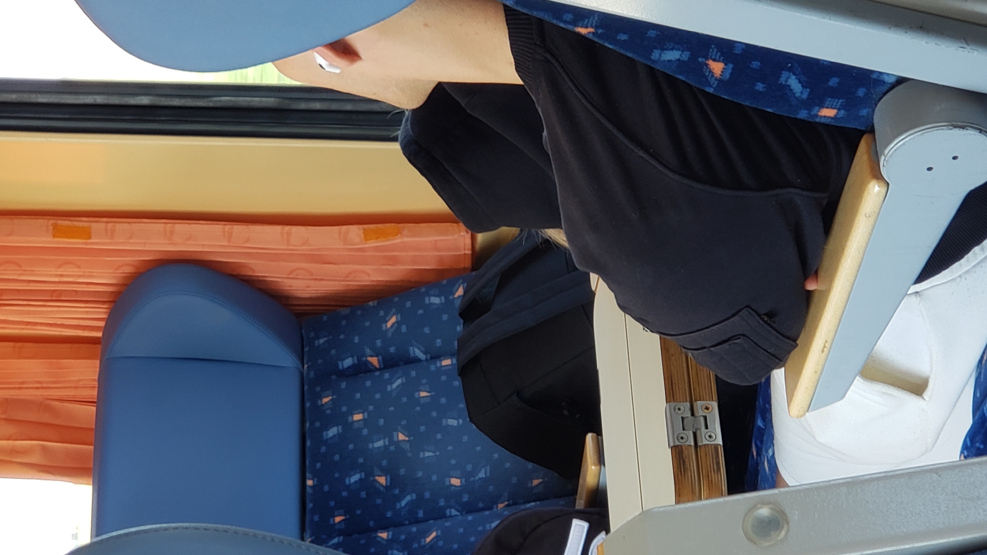 The image shows the interior of what appears to be a bus or train carriage. The scene is dominated by blue seats with an orange and blue patterned fabric. In the foreground, a person is partially visible, wearing dark clothing. Their head is tilted down, and their face is not clearly visible. A light-colored wooden tray table is extended, and the person's hand rests on it. To the left, a blue seatback is prominent, with orange curtains visible behind it. The overall lighting suggests daytime. The location is identified as Brunovce, Slovakia.