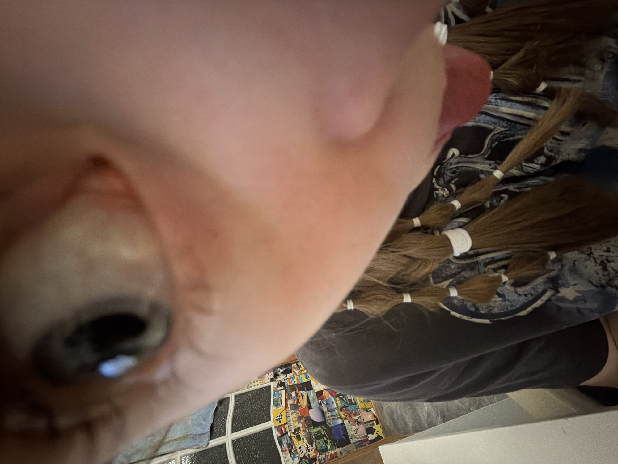 A close-up image features a person's face, predominantly focusing on their eye and cheek. The image is taken at a distorted, extreme angle. In the background, there are details of hair tied with white bands, suggesting braids or ponytails, and patterned fabric. Further in the background, there is a window with visible panes and a collage of what appears to be comic book or magazine clippings. The location, Perm, Russia, cannot be determined from the visual elements in the image.