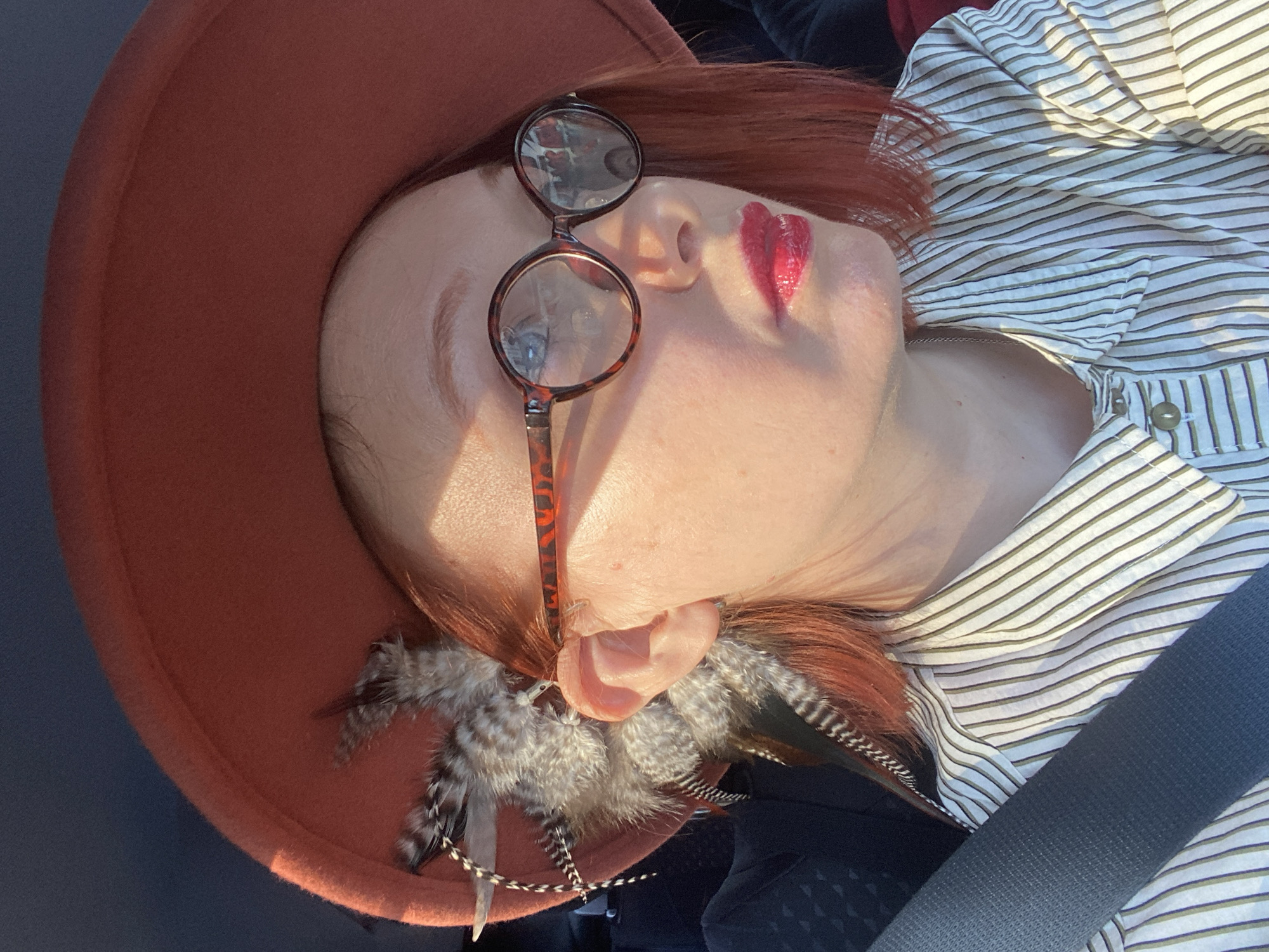 This close-up portrait, taken in Yekaterinburg, Russia, features a person, likely female, with reddish-brown hair, positioned with their head tilted to the right. They are wearing a wide-brimmed, reddish-orange felt hat. A pair of tortoise-shell-framed round glasses rests on the bridge of their nose, with the lenses reflecting light and surroundings. Their upper lip is distinctively painted with bright red, heart-shaped lipstick.
The person is wearing a light-colored, possibly white, shirt with thin, horizontal olive-green stripes. Near their right ear, a cluster of decorative feathers in shades of grey, black, and white with mottled patterns is visible, potentially part of an earring or hat embellishment. Strong, directional sunlight illuminates their face, hat, and shirt, casting warm highlights and soft shadows, indicating a sunny daytime setting.
The subject's eyes appear closed or are looking downwards, contributing to a serene or contemplative mood. A dark seatbelt is prominently visible across the lower right side of their shirt, suggesting they are seated in a vehicle, such as a car. The background is dark and out of focus, providing little specific environmental context beyond the immediate setting. No visible text is present in the image.