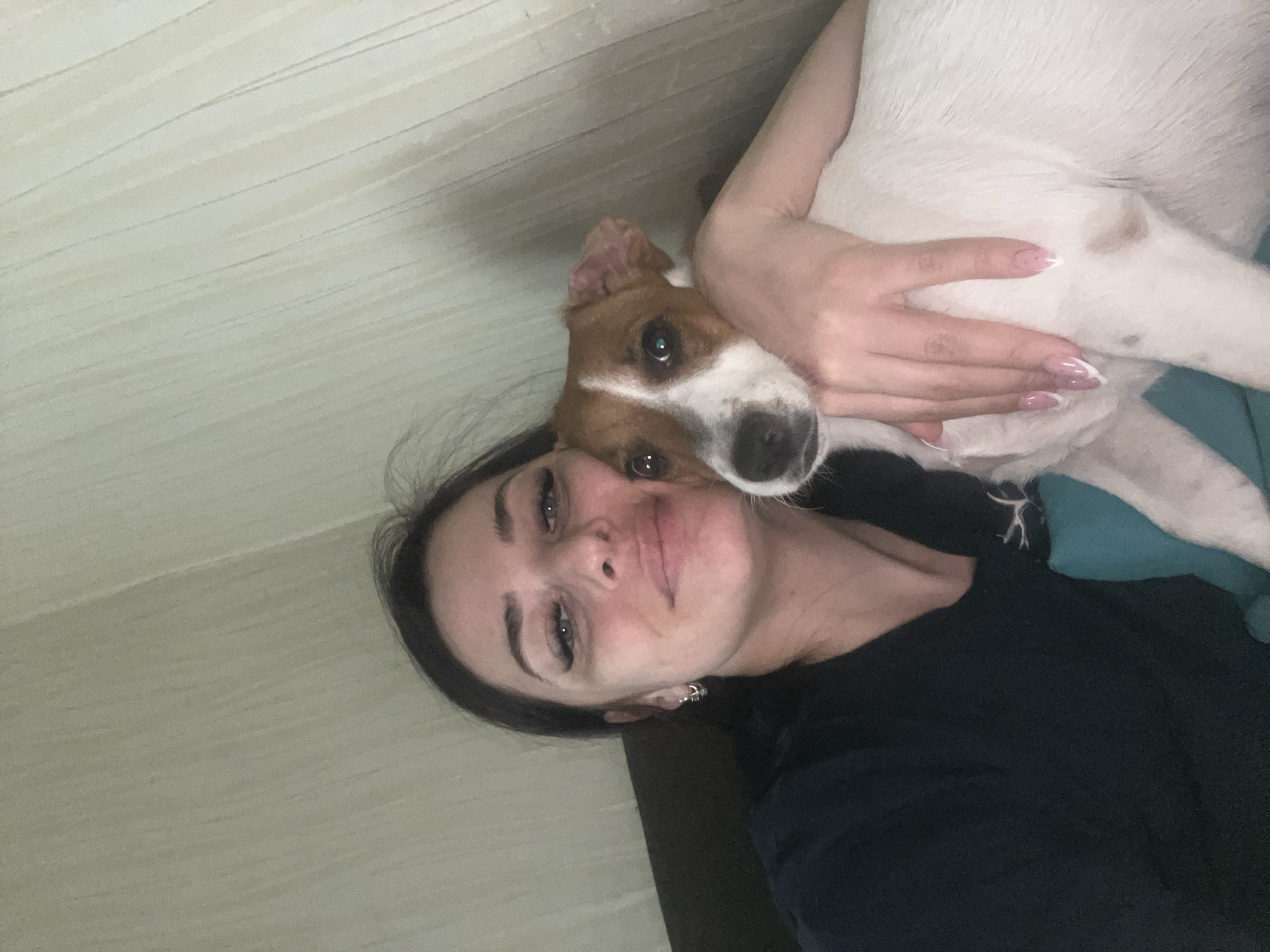 A horizontally oriented selfie captures a woman embracing a dog in an indoor setting. The young woman has dark hair and prominent eyelashes, with a soft, slight smile indicating affection. She wears a dark top, and her hand, adorned with a light pink and white French manicure, gently rests on the dog.
The dog, a small to medium-sized breed with brown and white fur, possibly a Jack Russell Terrier, is snuggled close to the woman, with its head resting near her cheek and shoulder. Both subjects look towards the camera, conveying a calm and affectionate mood.
The background features a light-colored wall with faint horizontal lines, suggesting wallpaper or paneling in a home environment. A hint of teal or turquoise fabric, possibly a blanket, is visible. The lighting is soft and diffuse. No specific time of day or external weather conditions can be discerned. There is no visible text in the image. The location is Chaikovsky, Russia.
