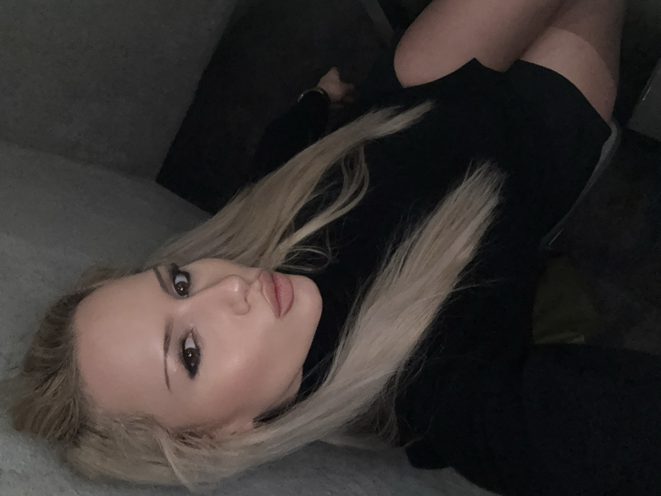An individual with long, straight blonde hair and prominent facial makeup, including dark eyeliner and lipstick, is depicted in a dimly lit indoor setting. The person, appearing female, is looking directly at the camera with a "pout" expression and is wearing a black long-sleeved turtleneck top or dress. The garment appears short, revealing the upper thighs, where a distinct tan line or hosiery is visible on the right leg. The background consists of a light grey, textured wall to the left of the subject and a dark, indistinct area to the right, possibly a corner or dark furniture. No specific identifying features of a location within Sofia, Bulgaria, are discernable from the visual information provided in the image.