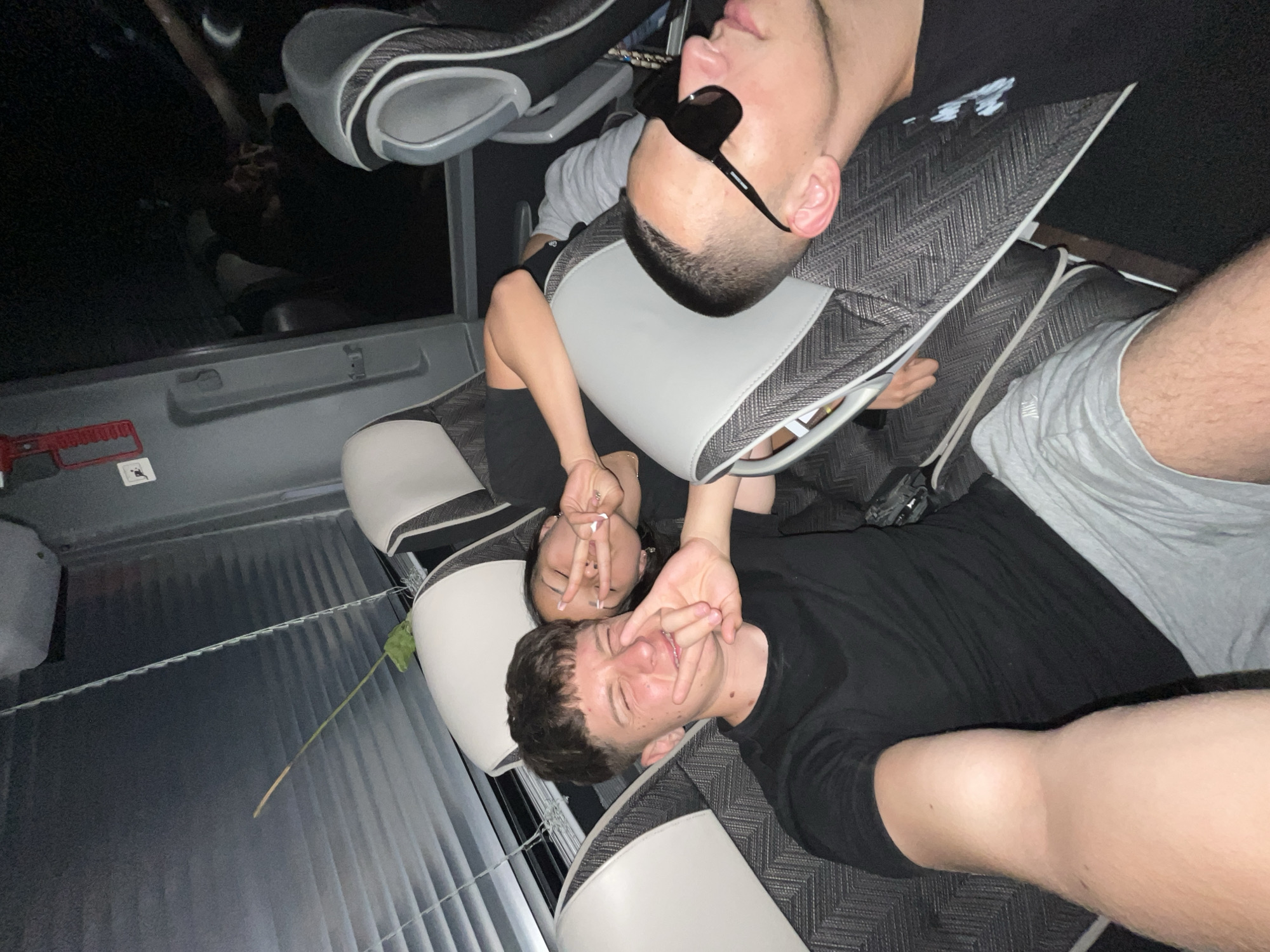 The image shows three people lying on a row of gray and white seats. One person wears sunglasses. Two people make peace signs with their hands. The background shows a window with closed blinds. There is no information to determine the city or the country.