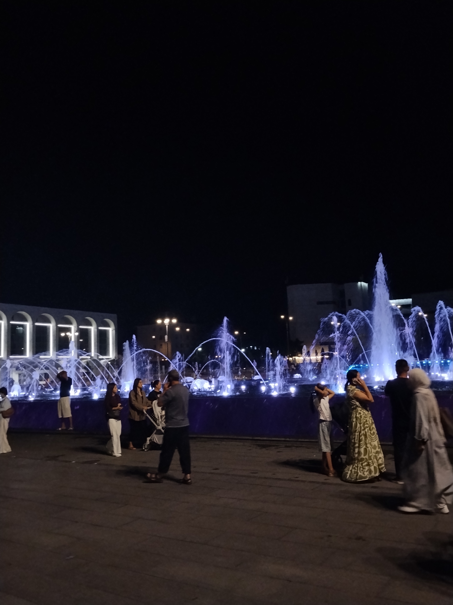A night-time outdoor scene depicts a group of people gathered near a large, illuminated fountain in Bishkek City, Kyrgyzstan. The fountain features multiple jets of water illuminated by blue lights, creating a dynamic display against the dark sky. In the background, a modern building with arched windows, also lit with white lights, suggests a public or commercial space.
Several individuals are visible in the foreground and mid-ground. On the left, a person stands near the fountain. To the right of the fountain, a man in a dark shirt and pants is seen from the back, facing the water. Beside him, a woman in a patterned dress is visible, and next to her, a person in a light-colored hooded garment is present. Further to the left, near the building, a group of people are standing and interacting. One person, wearing a white top and light-colored skirt, is facing away from the camera. Another individual, dressed in a maroon top and white pants, is looking towards the fountain.
The ground is paved, and shadows are cast by the people and the fountain's lights. The overall atmosphere appears to be one of leisure and social gathering.