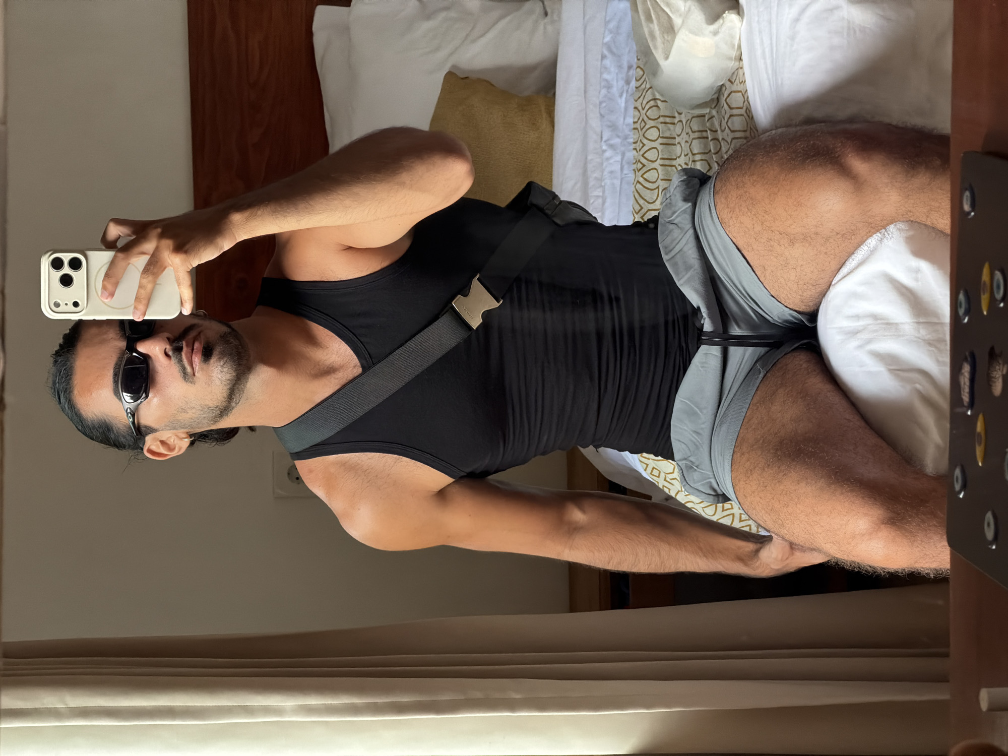 A man is captured taking a mirror selfie in what appears to be a bedroom or hotel room, with the user-provided context of North Kuta, Indonesia. He is positioned with his torso and legs visible, holding a white smartphone with a distinctive triple-lens camera array and MagSafe ring in his right hand.
The man has dark hair, styled away from his face, and a beard or stubble. He is wearing dark, wrap-around sunglasses. His attire consists of a black tank top and grey shorts. A black cross-body bag with a prominent gold-colored buckle, featuring indistinct text, is worn over his shoulder. His physique appears muscular, with visible arm and leg definition.
The setting includes a bed in the background, adorned with white pillows and a patterned blanket featuring a gold and white geometric design. A wooden headboard is partially visible. On the right side of the image, there's a dark piece of furniture or an object with several circular indentations. The lighting suggests it is daytime, with natural light subtly illuminating the room, possibly through a nearby window obscured by white curtains or blinds visible in the lower part of the image. The man's expression is neutral, consistent with posing for a photograph.