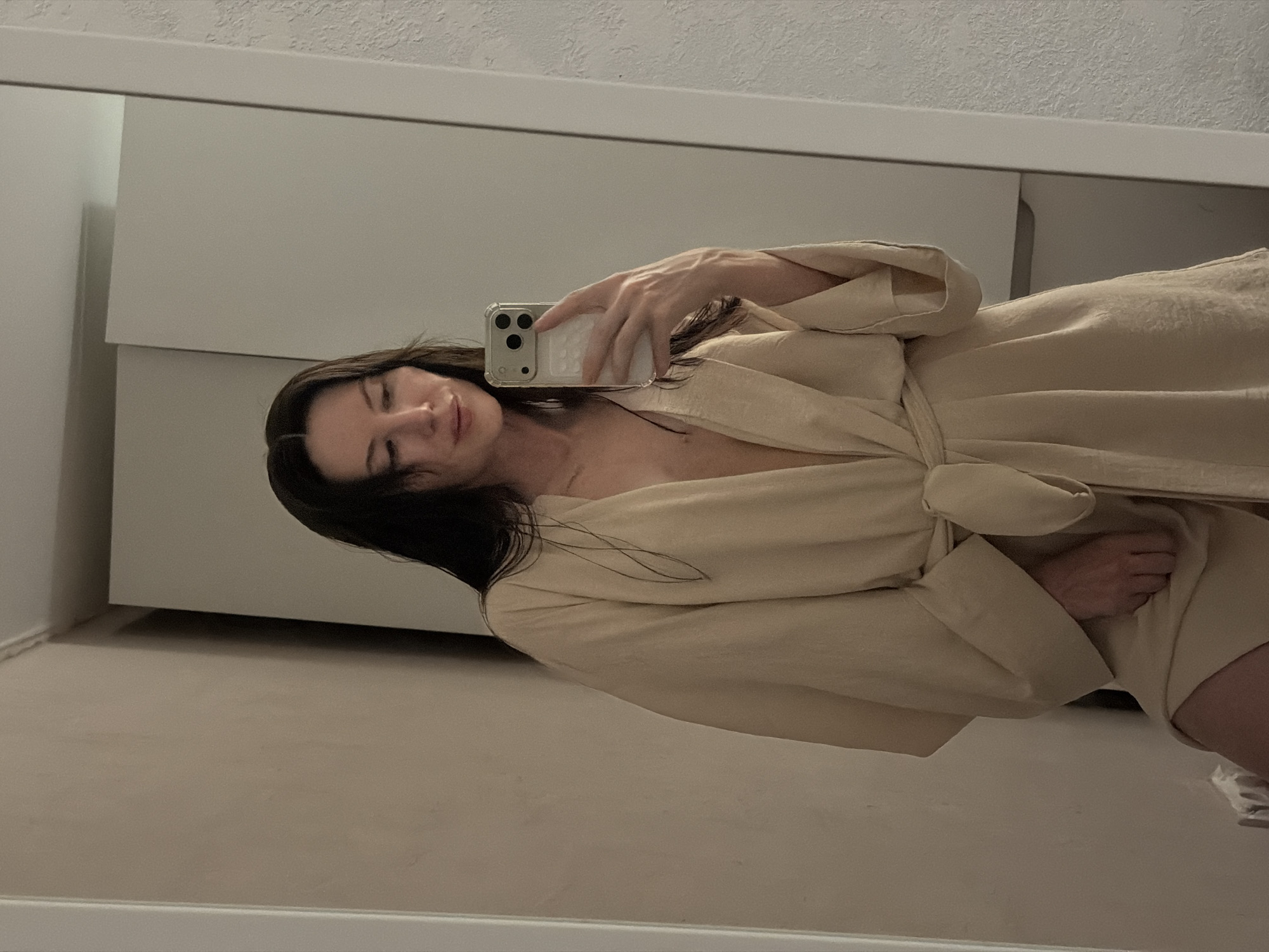 A woman in a beige robe holds an iPhone in her right hand, taking a selfie in front of a mirror. Behind her is a white wall and a white cabinet. The location is likely indoors. There is no evidence to suggest that the location is in Perm, Russia.