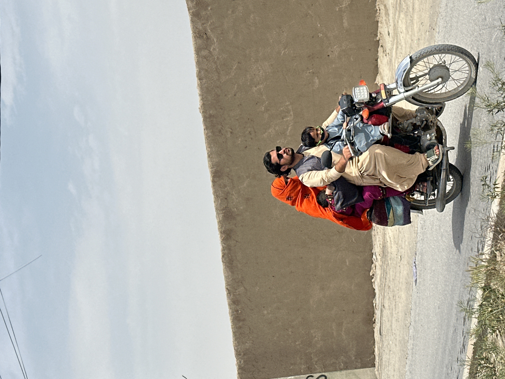 In an outdoor shot from Unknown, Unknown, a motorcycle carries five people. The foremost rider wears sunglasses and tan clothing. Two more individuals are between him and the driver, all tightly packed. At the rear is a person with an orange shawl. The motorcycle is on a paved road next to a light tan wall. The sky is visible.