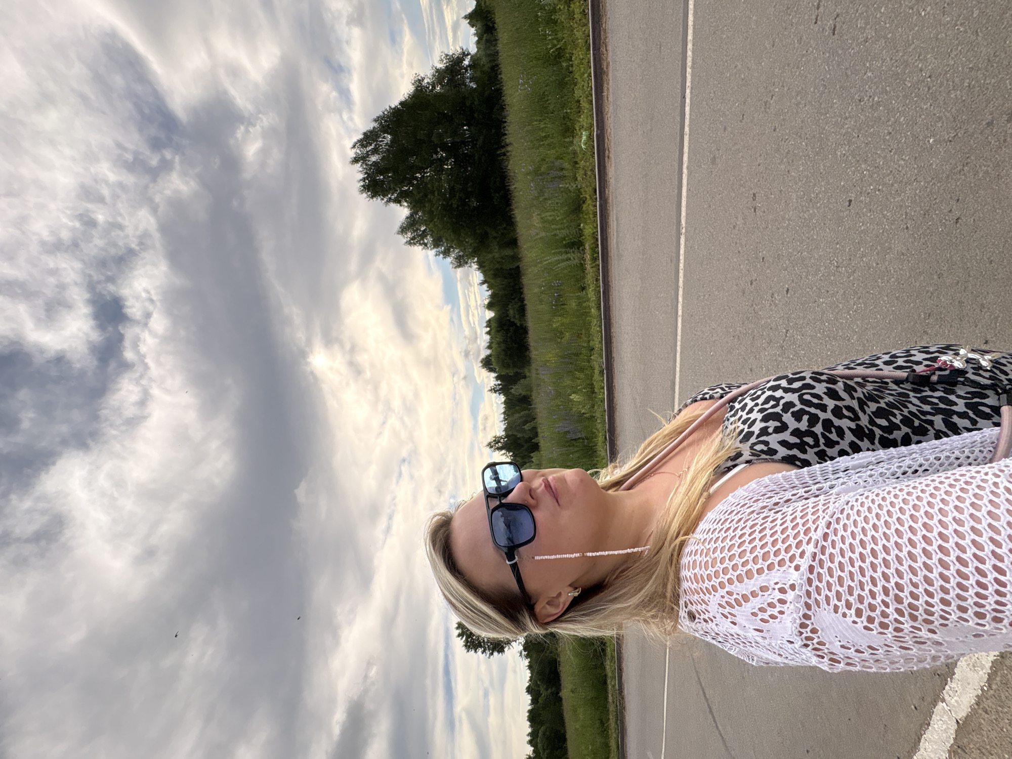 A blonde woman in her 30s or 40s, wearing sunglasses, a white lace cover-up, and a leopard-print dress, is standing on the side of a road. She is looking up and to the right, with a slight smile. The background consists of a field of green grass and flowers, with trees in the distance and a cloudy sky overhead. The image appears to be a selfie taken in a rural or suburban area, possibly on the outskirts of Марковский, Russia, given the presence of vegetation and a road. The lighting suggests it is daytime, with soft, diffused light coming from the sky.