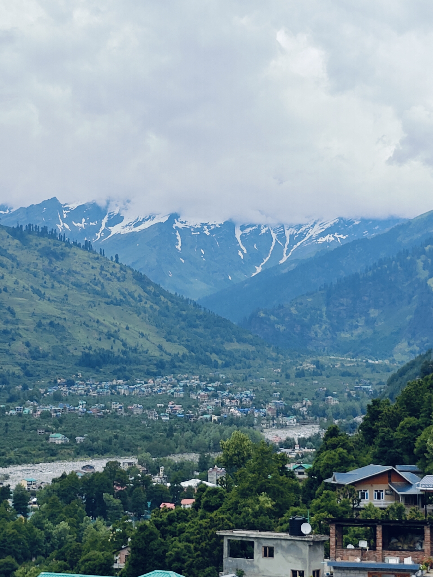 This image depicts a mountainous landscape in Vashisht, India. In the foreground, several buildings with various roofing materials are visible, nestled amongst dense green trees. One prominent building features a blue roof and a brown facade with white-trimmed windows. Further into the scene, a town or village with numerous buildings, many with blue and teal roofs, stretches across a valley floor. A river or rocky streambed is visible in the middle ground, separating the lower-lying town from the steep, green mountainside. The mountains themselves are covered in vegetation, with patches of snow and ice visible on the higher peaks. The sky is overcast with thick, grey clouds, partially obscuring the mountain tops. The overall scene conveys a sense of a remote, mountainous settlement.
