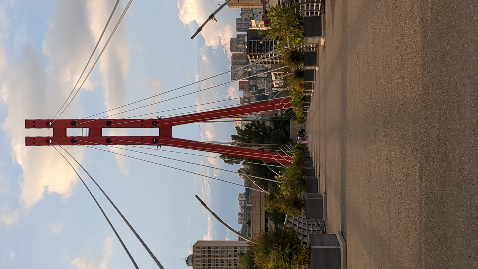 The image shows a pedestrian bridge in Tbilisi, Georgia, characterized by its prominent red A-frame pylon and a network of steel cables supporting the bridge deck. The perspective is from the bridge's pathway, looking forward towards the red pylon, which is silhouetted against a partly cloudy sky. Several people are visible walking on the bridge in the distance. Along the sides of the pathway are rows of large, dark rectangular planters containing lush green foliage. In the background, the cityscape of Tbilisi unfolds, with a mix of modern and older buildings, including a tall, light-colored building with a dome-like structure on its left. The overall scene suggests a pleasant day, possibly late afternoon or early evening, given the quality of light.