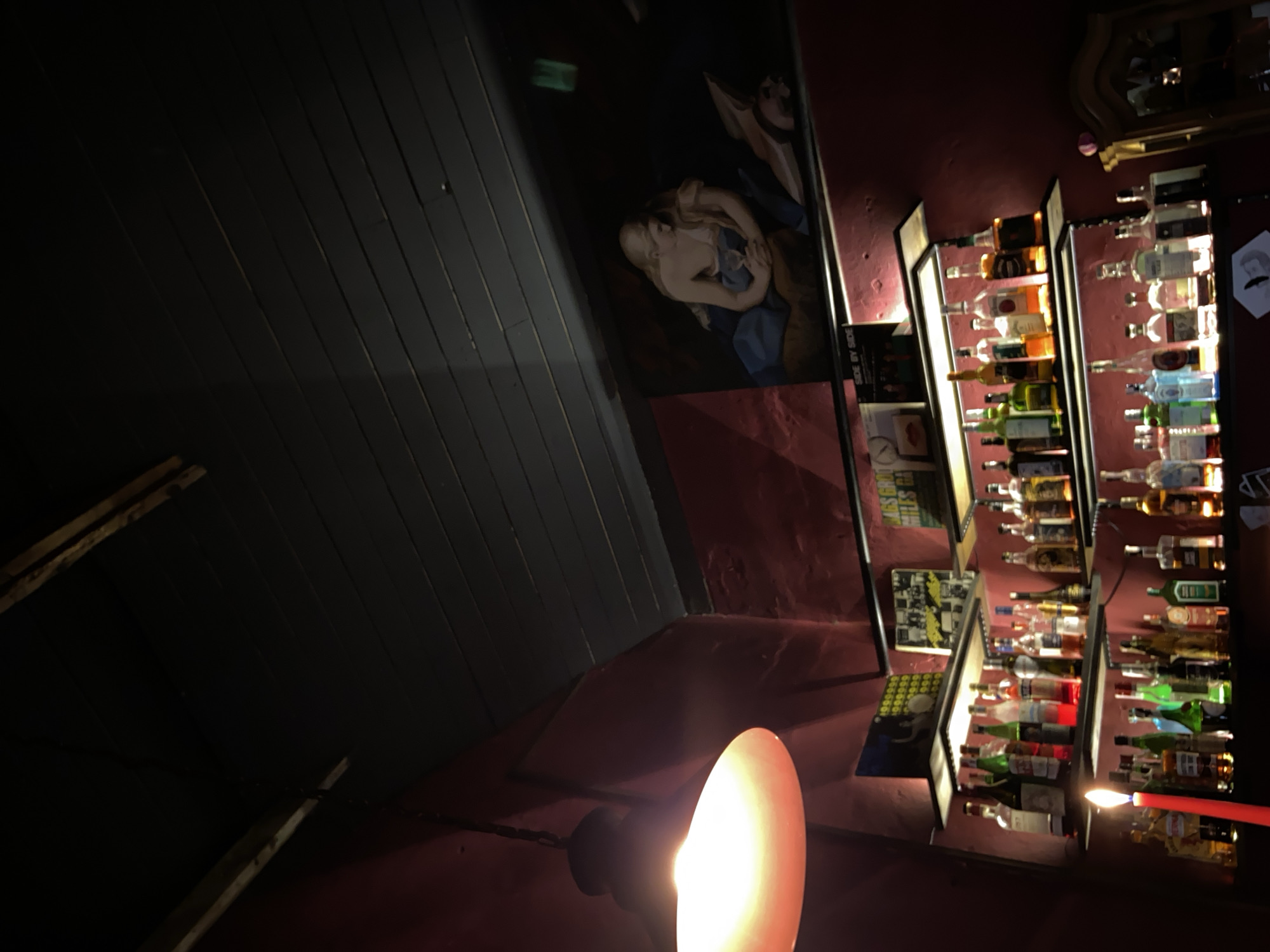 The image depicts the interior of a dimly lit bar, likely in Brest, Belarus. The scene is dominated by shelves laden with various bottles of alcohol. On the wall above the shelves hangs a painting of a partially clothed woman. A prominent light fixture with a large, glowing orange shade hangs from the ceiling on the left side of the frame. In the foreground, a tall red candle with a lit flame is positioned on a lower shelf, casting a warm glow. The overall atmosphere is dark and intimate, suggesting a nocturnal setting within a establishment serving beverages.