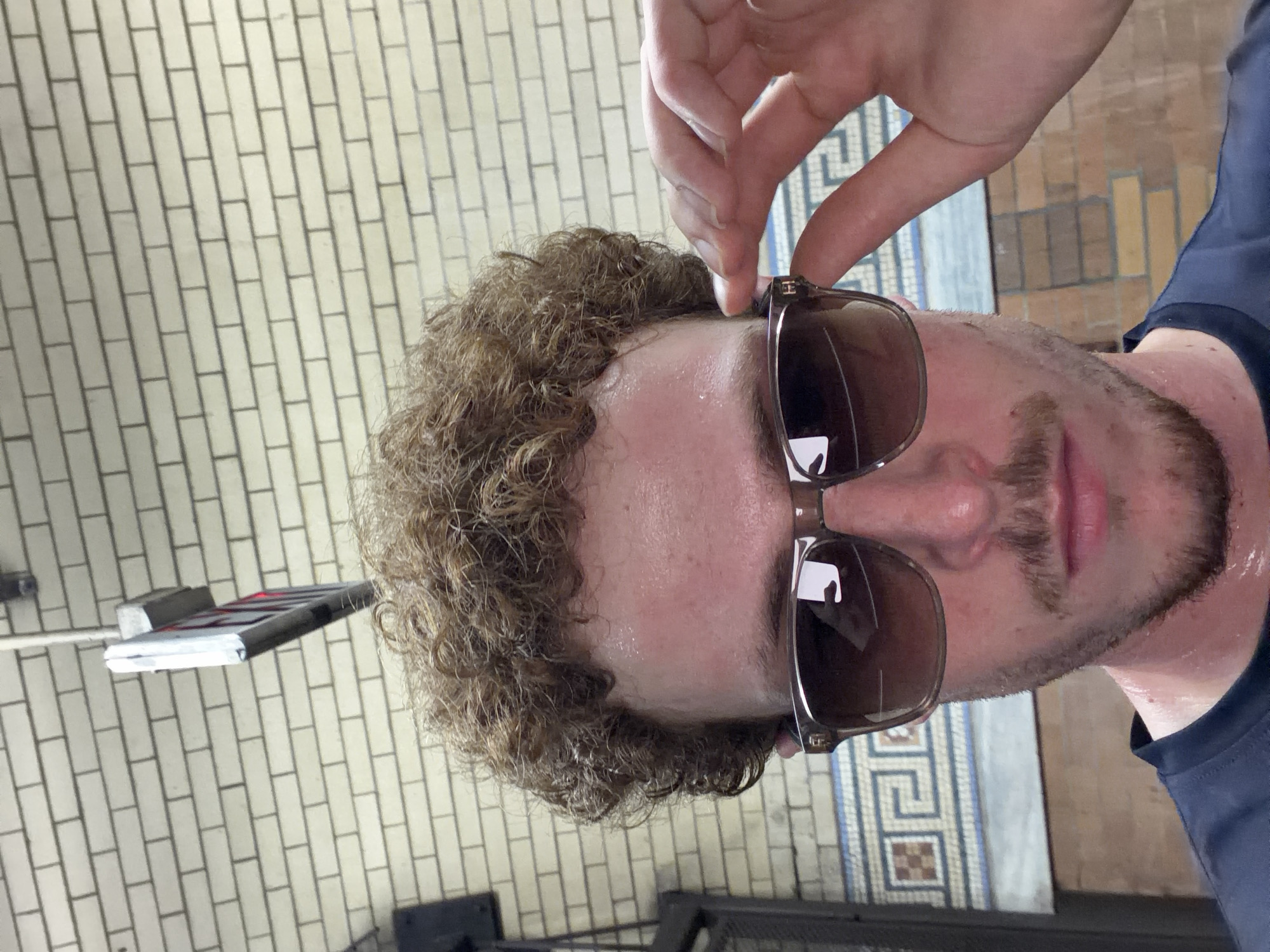 A man with curly brown hair and a mustache, wearing sunglasses, is standing in a New York City subway station. He is holding the sunglasses with his right hand. The subway station has white brick walls, and a floor with a mosaic tile pattern. There is a black metal fence behind the man. A sign hangs from the ceiling. The man appears to be sweating.