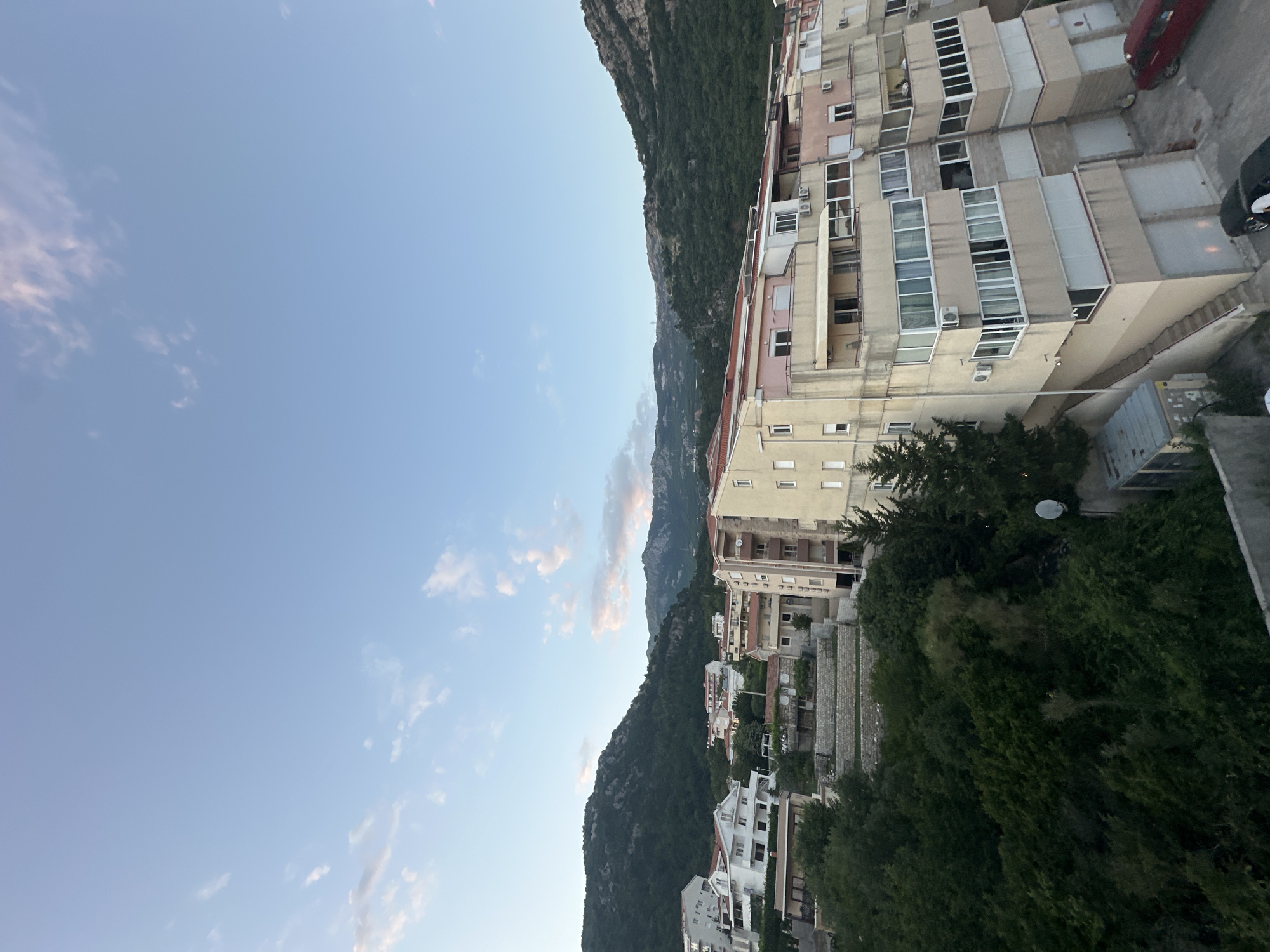 The image captures a vertical shot of a cityscape nestled against a mountain, likely in Budva, Montenegro. The sky is a clear blue with a few scattered clouds. On the right, multi-story residential buildings are visible. The buildings appear to be constructed from concrete with white balconies and windows. The exterior of the buildings are painted in light neutral colors. Adjacent to the buildings, the mountain is covered with dense vegetation. Closer to the base of the mountain, houses are dispersed among the trees. A red car is parked near the top right corner of the image.