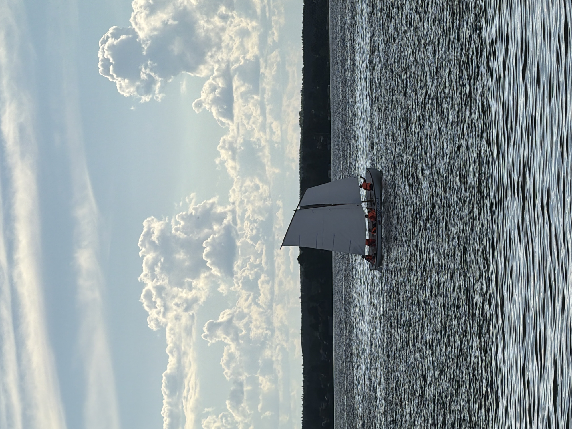 The image depicts a sailboat with multiple people on board sailing on a body of water. The background shows a tree line and a sky filled with white, fluffy clouds. The water surface is rippled, reflecting the sky. The people in the sailboat are wearing orange life vests. Without additional data or sources, it is impossible to confirm the image's location in Votkinsk, Russia or to confirm the accuracy of a location.
