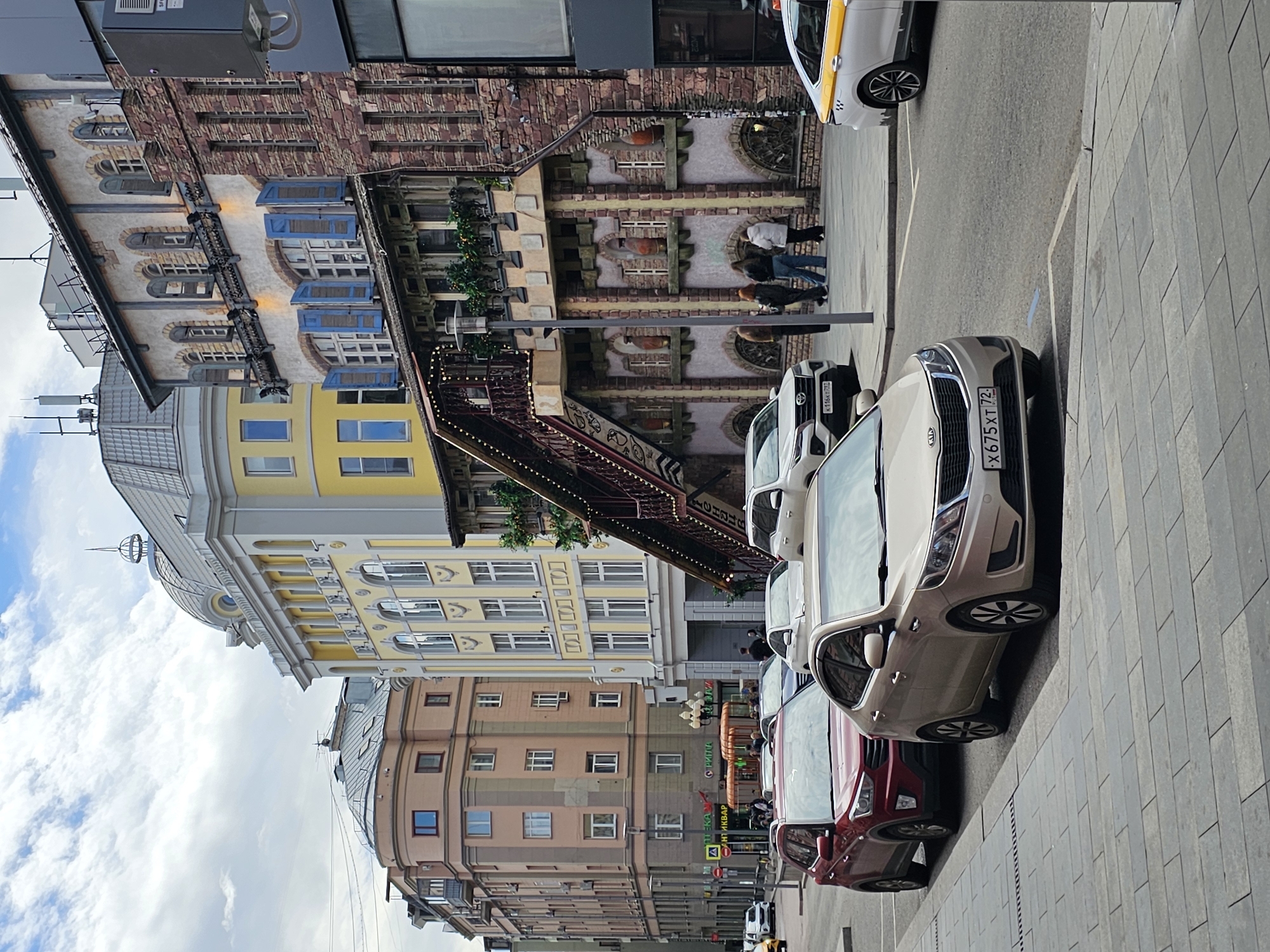 The image captures a street scene in Moscow, Russia, notable for its diverse architectural styles. From the street level upwards, a row of parked cars is visible, including a white car and a red car. Further up, the architecture ranges from a building with brown and beige hues to a structure with a yellow façade and a domed top. In the background, a brick building displays an elaborate design with wooden accents and multiple windows. Pedestrians are seen walking in front of this building. The sky is partly covered with clouds. Overall, the scene showcases a blend of historical architecture and modern urban life.