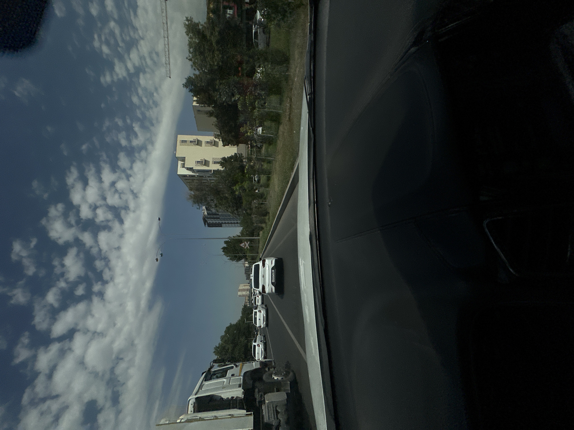 This is a forward-facing, eye-level shot from inside a vehicle on a multi-lane paved road in Tashkent, Uzbekistan. The sky is blue with scattered white cumulus clouds, and the sun is bright. A large white truck is in the left lane, followed by several white cars driving in the same direction. Buildings and trees line the right side of the road, with a construction crane visible in the distance. The foreground shows the dashboard and windshield of the vehicle.