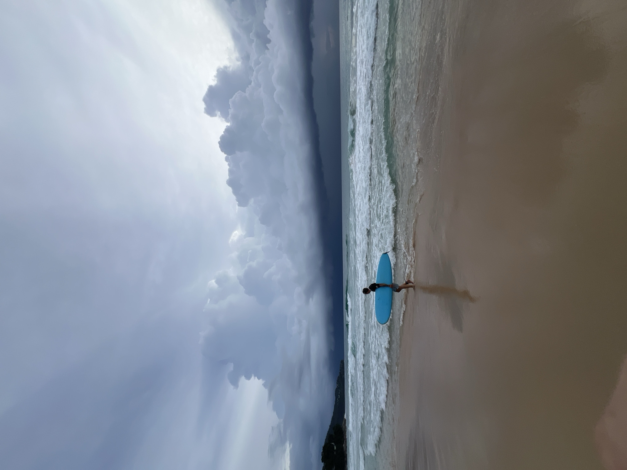 The image depicts a person standing on a sandy beach holding a blue surfboard. The person is wearing a dark top and light-colored shorts. They are positioned near the edge of the water, where gentle waves are breaking on the shore. The beach sand appears light brown and wet near the water's edge, gradually becoming drier and lighter further inland.
The sky is dramatic and overcast, with large, dark storm clouds dominating the left side of the frame. Rays of light are visible piercing through the clouds, suggesting a dynamic weather system. The ocean water is a greenish-blue, reflecting the cloudy sky. In the distance, a line of trees and possibly hills can be seen along the coastline.
The overall mood is one of anticipation or transition, possibly due to the approaching storm. The time of day is unclear due to the heavy cloud cover, but the lighting suggests it could be afternoon. The location cues and tropical vegetation hint at a beach in a warm climate, consistent with the provided location context of Choeng Thale, Thailand. There is no visible text in the image.