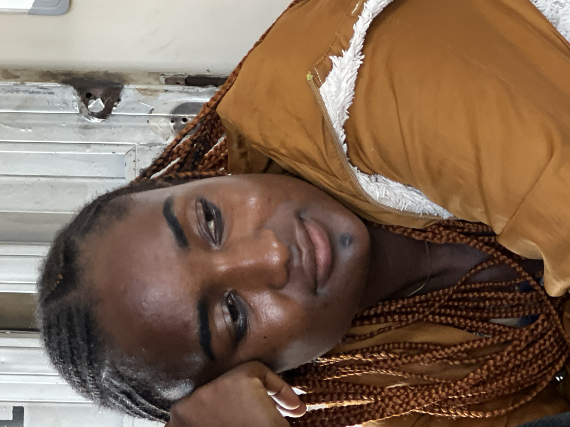 This image is a close-up portrait of a young Black woman with braided hair, wearing a brown jacket lined with white fur. She is leaning her head on her hand, looking away from the camera with a thoughtful expression. The background appears to be an indoor setting with a weathered, metallic door or partition. The lighting suggests an indoor environment, possibly during the day. The image does not contain any specific visual cues that would indicate its location within the city of Jos, Nigeria.