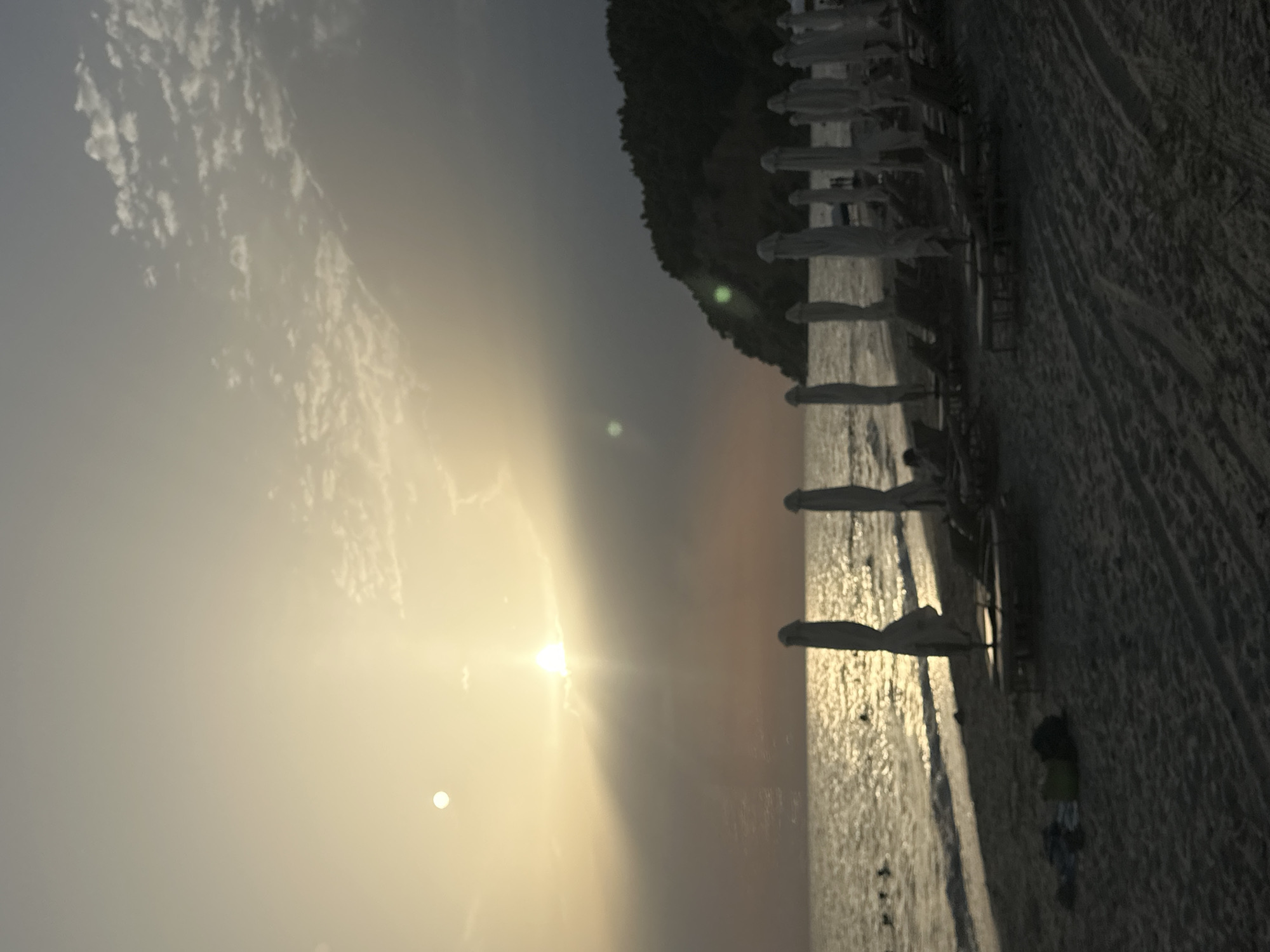 A beach scene at dusk in Kassandra Municipal Unit, Greece, features a body of water reflecting the setting sun, with waves gently lapping at the shore. Numerous closed beach umbrellas and empty lounge chairs are neatly arranged on the sandy beach, suggesting a quiet end to the day. A dense, tree-covered hill rises on the right side of the frame, silhouetted against the darkening sky. A few people are visible in the water in the distance, seemingly enjoying a last swim before sunset. The sky is a gradient of soft yellow and muted gray, with scattered clouds adding texture.