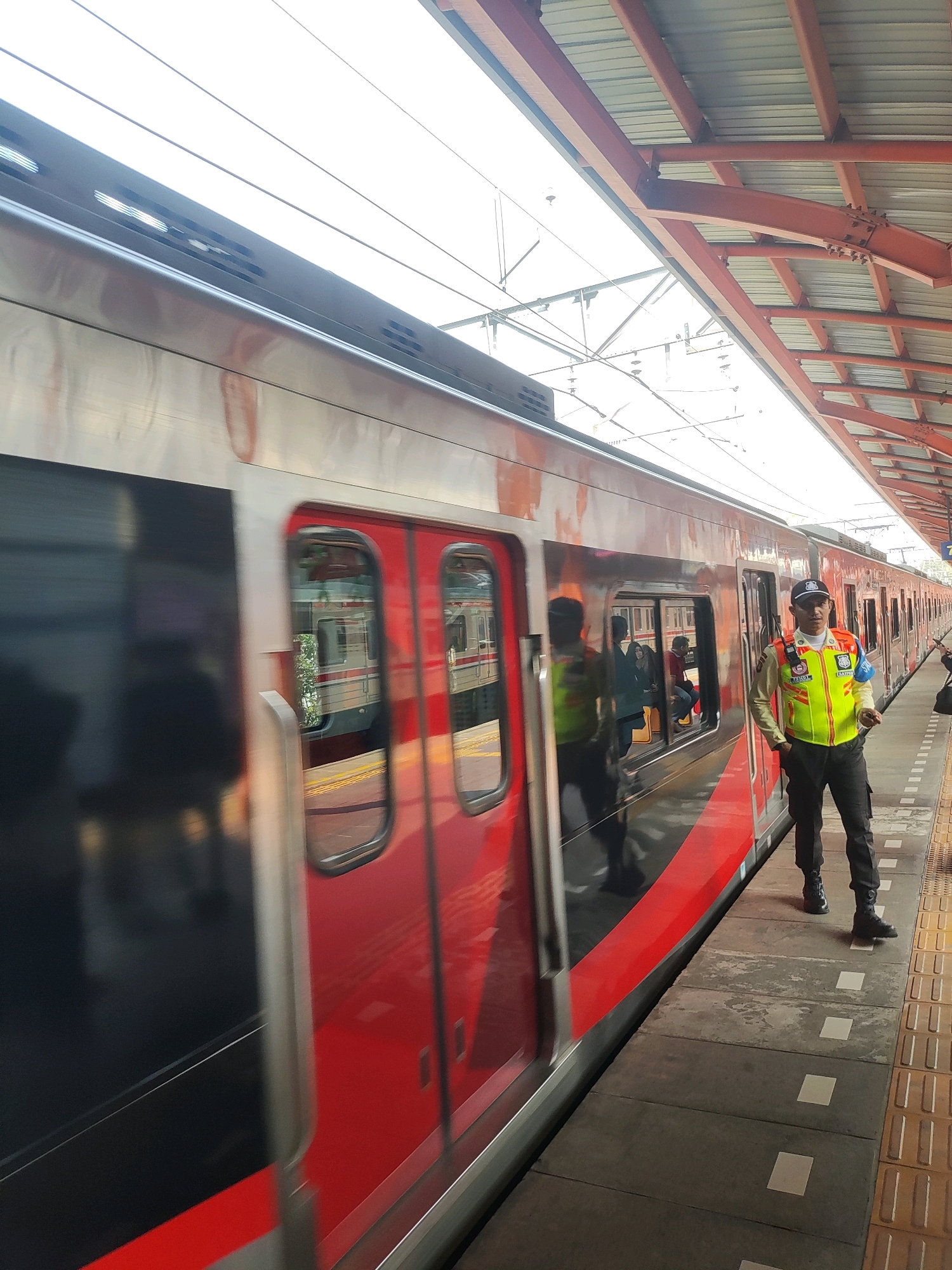 A man in a bright yellow safety vest, dark pants, and a black baseball cap stands on a train platform in Bekasi, Indonesia. He is facing towards a red and silver train that is either stopped or moving slowly. The train has dark windows and doors. The platform is made of textured yellow tiles with tactile paving. Above the platform, a red metal canopy supported by beams covers the train and tracks. The sky is bright and appears to be overcast or hazy. In the background, the train extends down the platform, and there are reflections of people inside the train's windows.