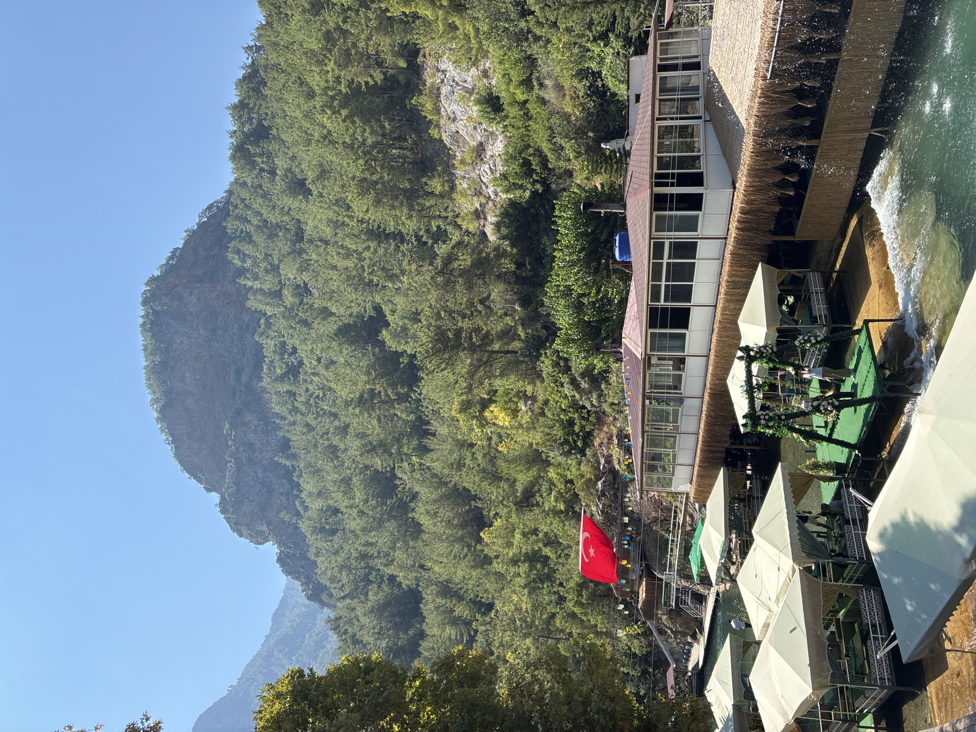 The image depicts a scenic outdoor restaurant with overwater seating in Alanya, Turkey. The restaurant features multiple elevated platforms with white canopies, providing shade for diners. Some platforms are directly over the clear, shallow water, while others are on the edge of the water. There are tables and seating arranged on these platforms. A swing decorated with white flowers is also visible.
In the background, a large, lush green mountain covered in pine trees dominates the landscape. A Turkish flag is prominently displayed on a flagpole on a raised section of the restaurant. The water flowing through the restaurant area appears to be a natural water source, possibly a river or stream, with small waterfalls adding to the ambiance. The sky is a clear, bright blue. The architecture of the main restaurant building, which has a red-tiled roof and large windows, is visible behind the overwater seating. There are a few people present, casually dressed, seemingly enjoying the atmosphere. The overall scene conveys a relaxed and picturesque dining experience in a natural setting.