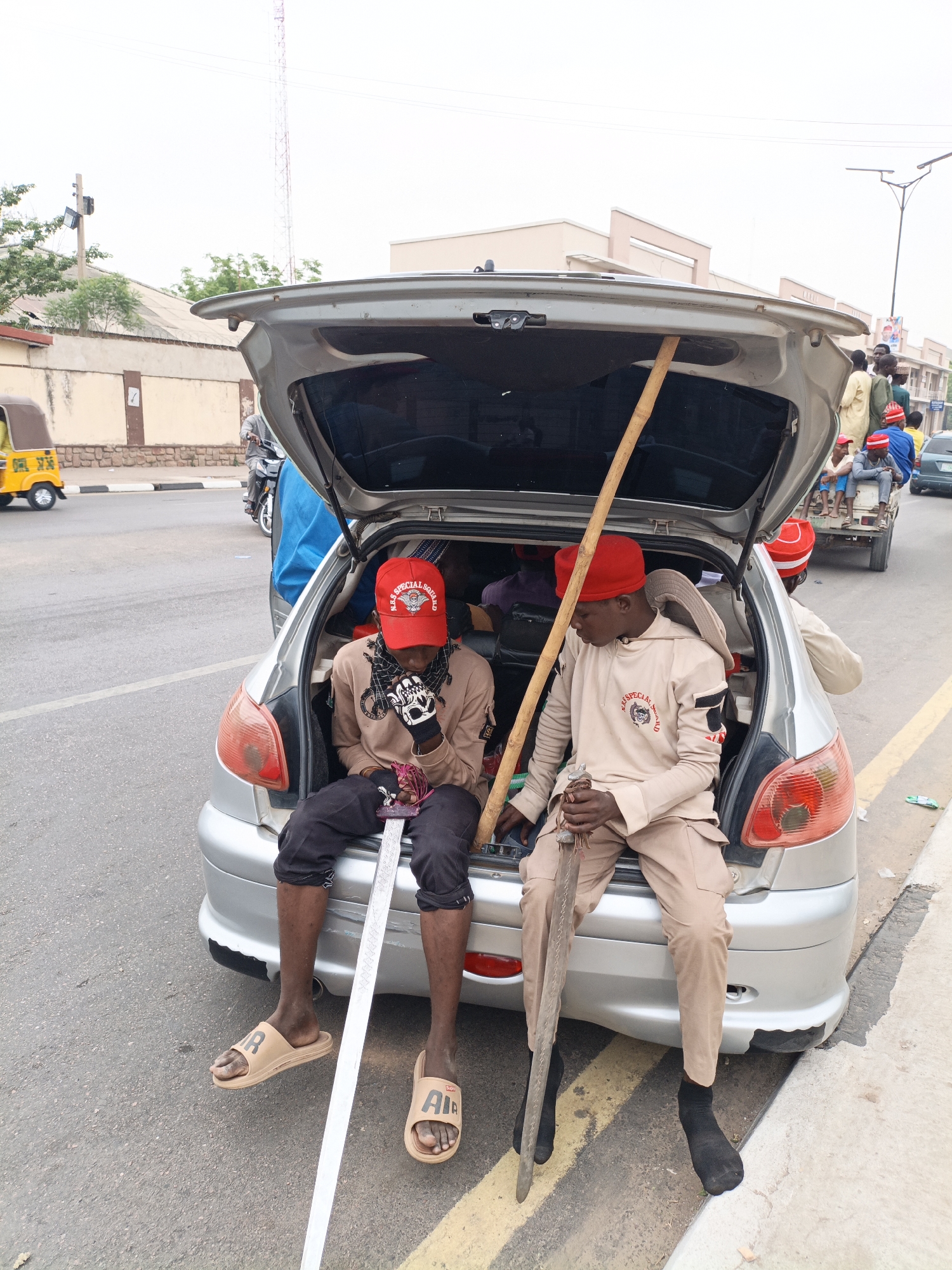 Two young men are sitting in the open hatchback of a silver compact car on a street in Tudun Wada, Nigeria. The man on the left wears a red cap, a brown shirt, black pants, and brown sandals. He is holding a long, flat metal object that resembles a sword. The man on the right is wearing a red cap, a tan uniform, and black socks. He is holding a brown, curved object that resembles a machete. A long wooden pole is leaning inside the car, between the two men. In the background, other people are sitting on a small trailer being towed by a car. A yellow three-wheeled vehicle is visible on the left side of the frame. The sky is overcast.