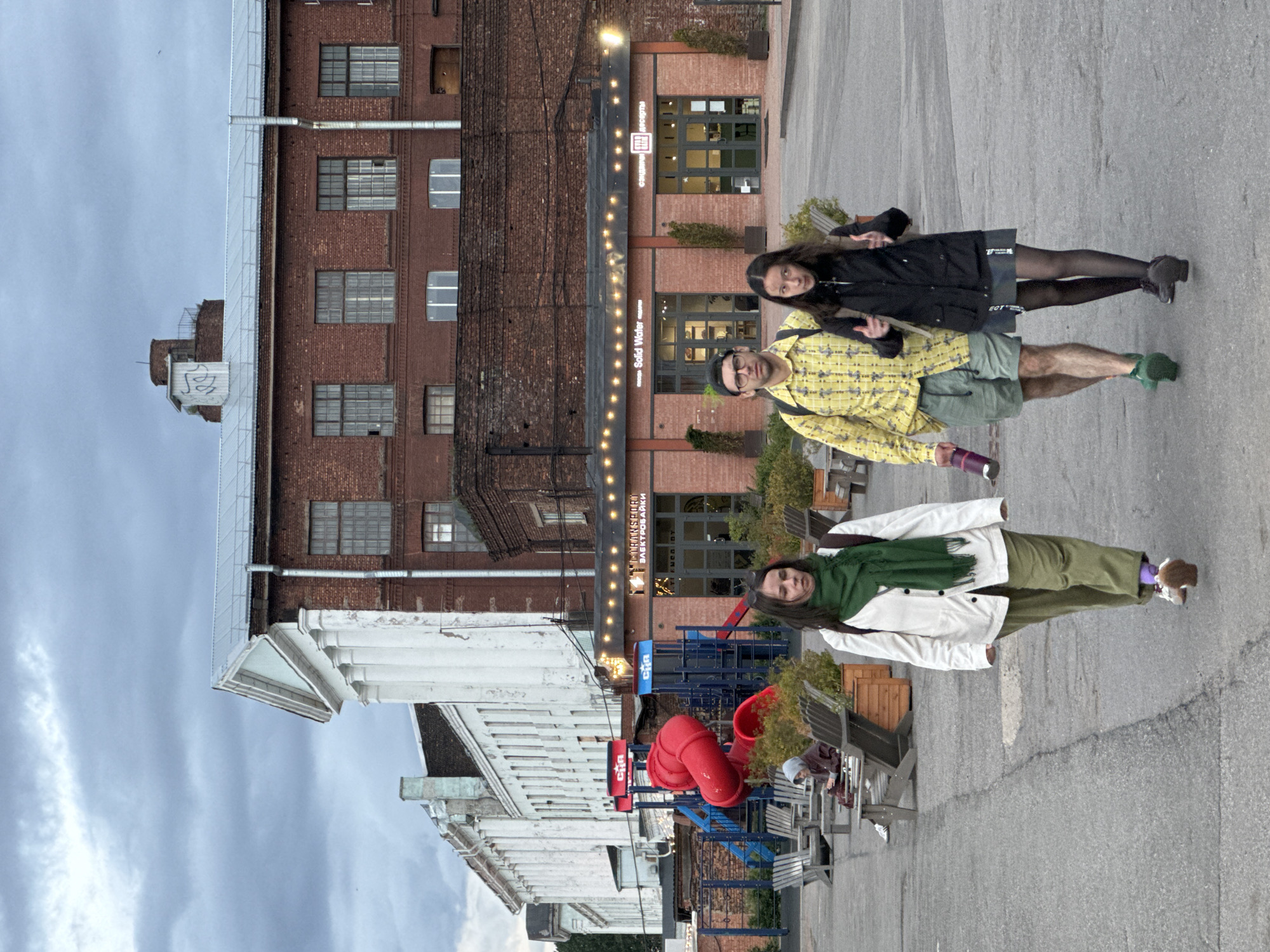 The image captures an outdoor scene in Saint Petersburg, Russia, likely in a renovated industrial or public space. In the background, a large, old brick building with a white classical facade is visible, adjacent to a newer brick structure with a pitched metal roof. The newer building features a row of windows and a storefront with a sign that reads "Solid Water" and Cyrillic text. Strings of lights are hung above the storefront.
In the mid-ground, a playground with a bright red slide and blue structural elements is situated in front of the buildings. Several wooden chairs are arranged in seating areas.
In the foreground, three individuals are walking towards the viewer on an asphalt paved area. From left to right, a woman with dark hair wears a white jacket, green scarf, and olive-colored pants. Next to her, a man with glasses and a cap wears a yellow plaid shirt, green shorts, and green shoes. He holds a dark water bottle. On the right, a woman with dark hair wears a black jacket, a gray skirt, and black tights. She gestures with her hands.
