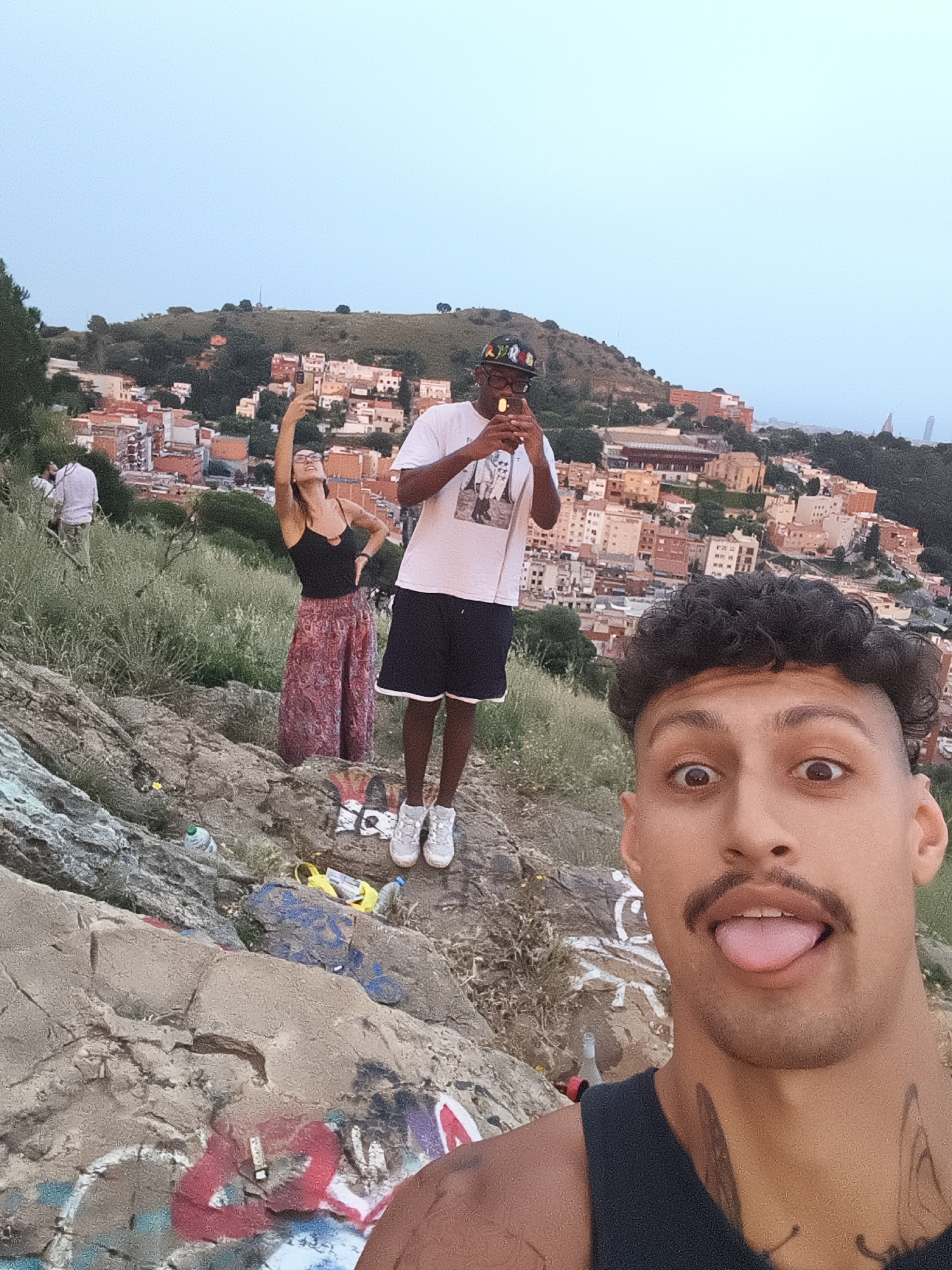 Three people are taking photos on a hillside overlooking the city of Barcelona, Spain. The foreground features a selfie of a man with dark curly hair, a mustache, and tattoos on his neck and shoulder, sticking his tongue out at the camera. Behind him and slightly to the left stands a woman in a black tank top and a long patterned skirt, holding up her phone to take a picture. To the woman's right is a man wearing a white t-shirt, black shorts, white shoes, and a colorful hat, also holding up his phone as if taking a photo or video. The hillside is rocky and has graffiti on some of the rocks. There is trash (bottles and bags) around their feet. The background shows a dense urban landscape with buildings clustered together, and a hill beyond them. The sky is overcast and light blue. A fourth person is present in the background, further left.