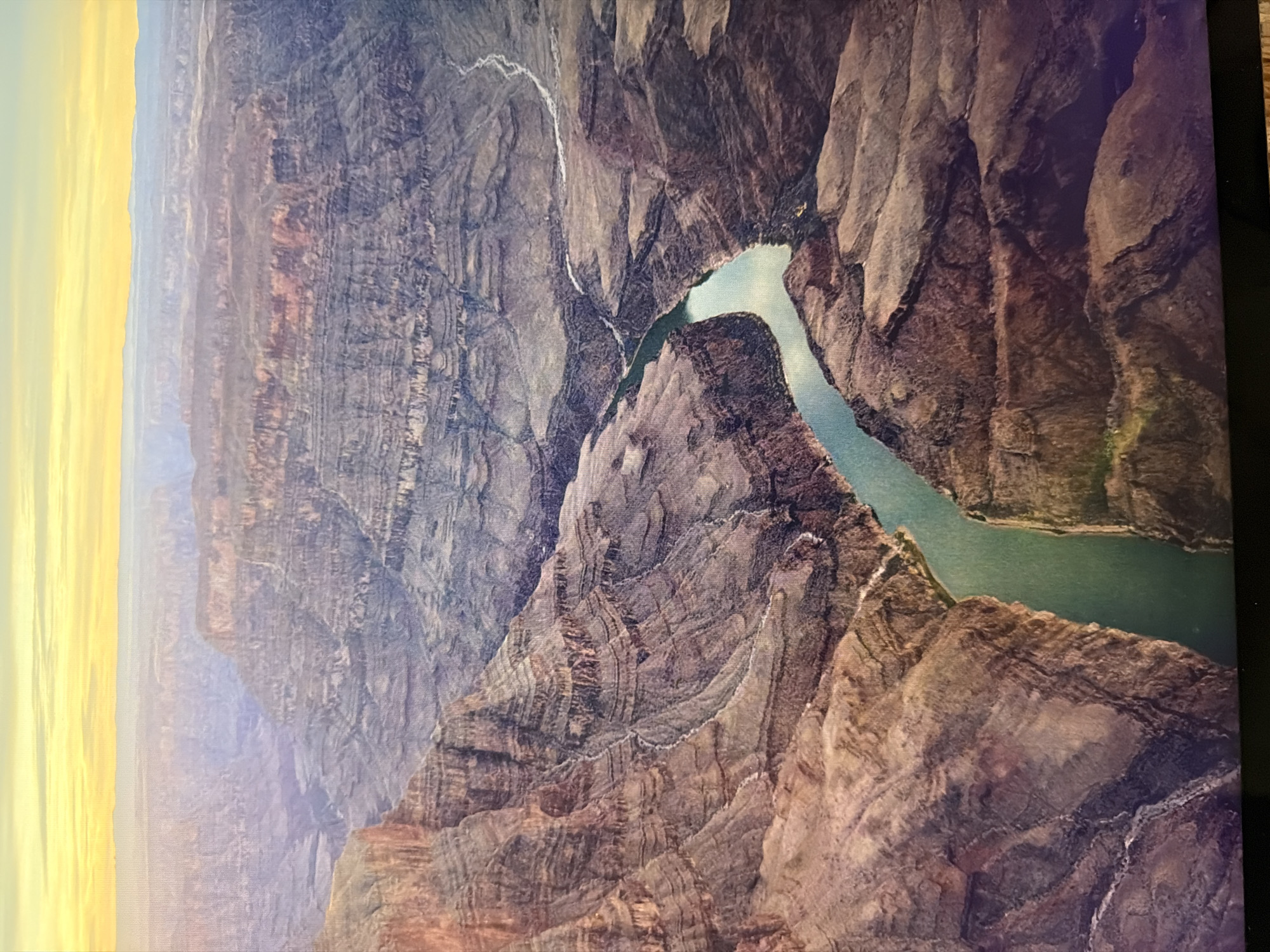 This is a drone shot aerial view of the Grand Canyon at sunset. The sky is a gradient of yellow and orange, with distant mountains silhouetted against it. The canyon walls are layered with shades of brown, red, and gray, showing the geological history of the area. A turquoise river winds through the bottom of the canyon, reflecting the sky. The scene is vast and serene, capturing the immense scale and natural beauty of the Grand Canyon. The image does not depict any scenes or locations within the city of Kyiv, Ukraine.