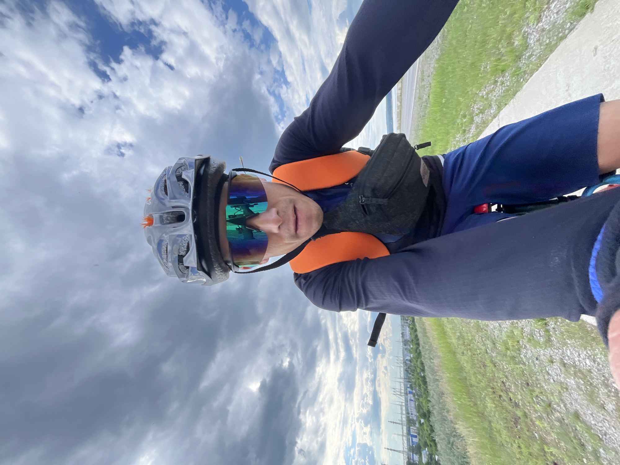 An outdoor, selfie-style, eye-level, wide-angle shot captures a cyclist wearing a helmet, mirrored sport sunglasses, and a dark long-sleeved shirt with an orange safety vest over it. The cyclist also wears a black fanny pack around their waist and dark blue shorts. The cyclist is on a bicycle, with a portion of the handlebars visible. The background shows a grassy embankment with a path or road, and in the distance, industrial structures with tall stacks are visible, likely within the city of Chaikovsky, Russia. The sky is cloudy with patches of blue. The cyclist's right arm is extended, holding what appears to be the camera or phone taking the picture.