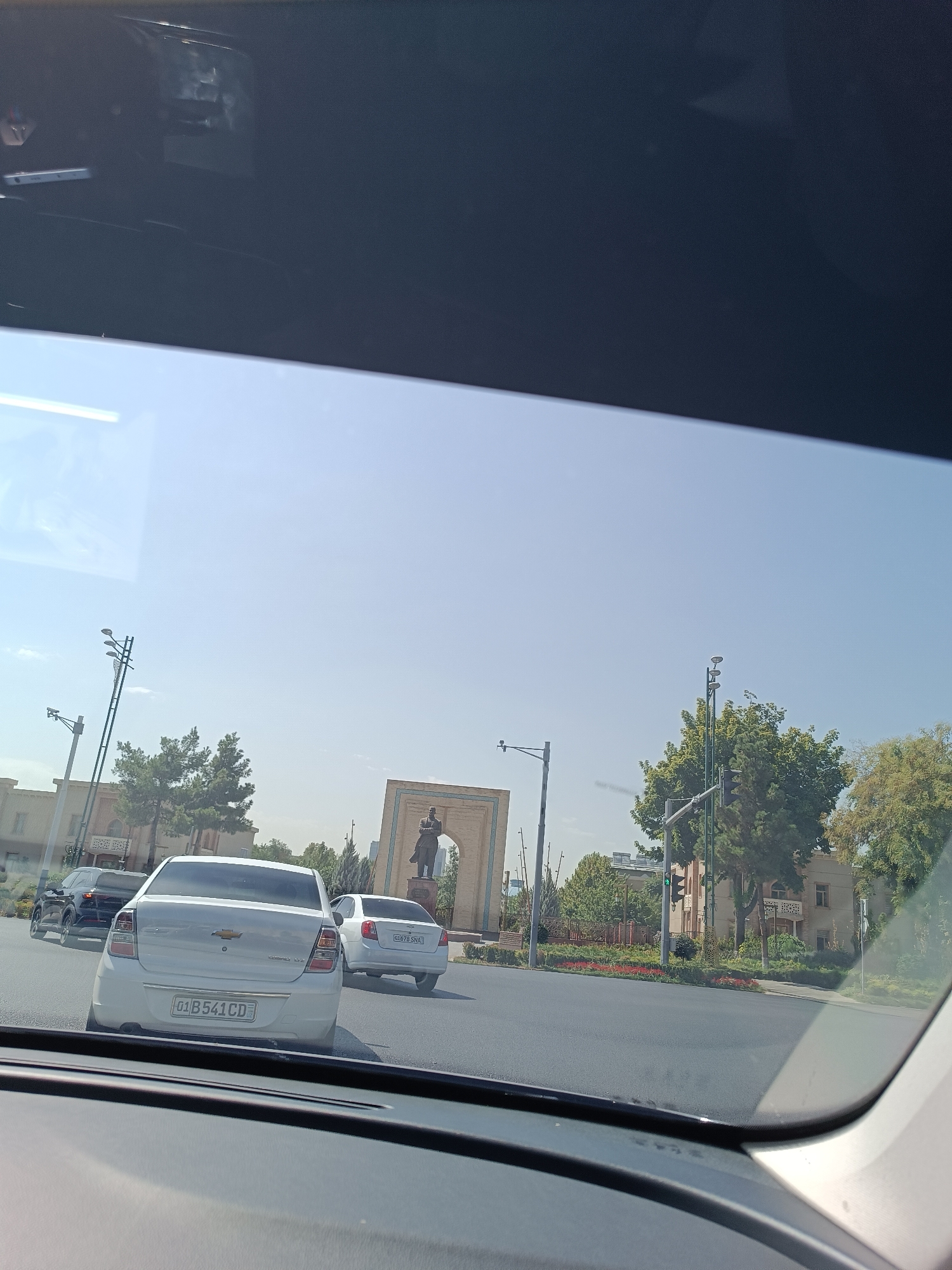 The image shows a view from inside a vehicle looking out through the windshield at a street scene in Tashkent, Uzbekistan. In the foreground, the dashboard of the vehicle is visible. The street is paved and appears to be an intersection. Several cars are on the road, including a white Chevrolet in the center foreground with a license plate reading "01 B 541 CD". To the left of the Chevrolet is a black SUV. To the right of the Chevrolet, another white car with a license plate reading "07 G79 SNA" is visible.
In the background, there is a statue of a man in traditional attire standing under an archway. Behind the statue, there are buildings with architectural features that suggest a historical or cultural significance. Trees line the street and surround the buildings, and there are traffic lights and streetlights visible. The sky is clear and blue. The overall impression is of a sunny day in a city with a blend of modern and traditional elements.