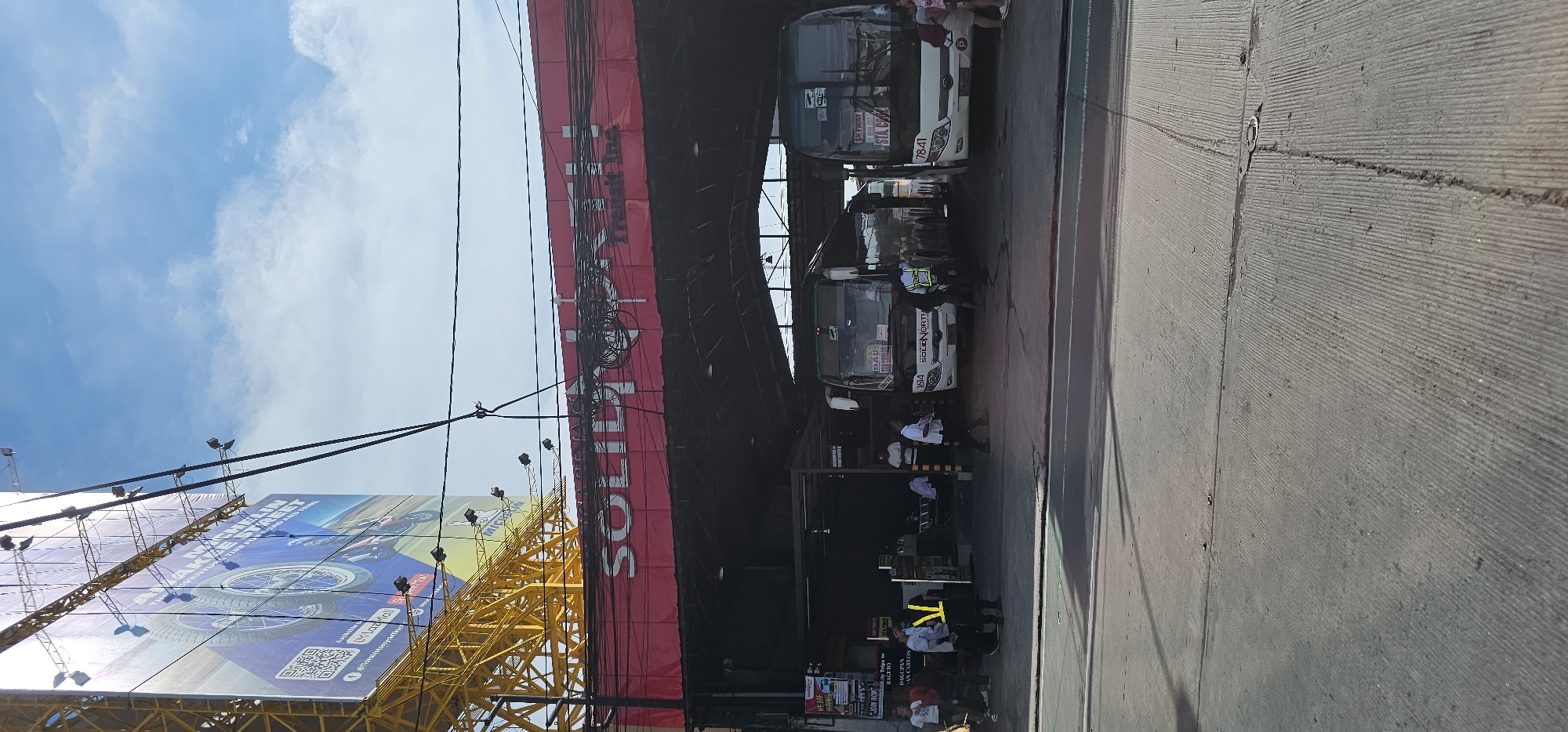 This is a medium-shot, eye-level photograph taken on a bright, cloudy day at a bus terminal in Quezon City, Philippines. The foreground shows a paved road with visible tire marks and light shadows. In the middle ground, a bus terminal with a red awning and "SOLIDARITY TRANSIT INC." signage is visible. Several white and dark-colored buses are parked under the awning. A few individuals, including men in white shirts and one in a yellow vest, are seen near the buses. Some of the buses have destination signs that read "BAGUIO" and "STA. CRUZ". A large billboard with a tire advertisement is prominently displayed on the left side of the frame, towering over the terminal. The background consists of a blue sky with scattered white clouds. The scene depicts a typical day at a public transportation hub, with buses preparing for or having completed their journeys.